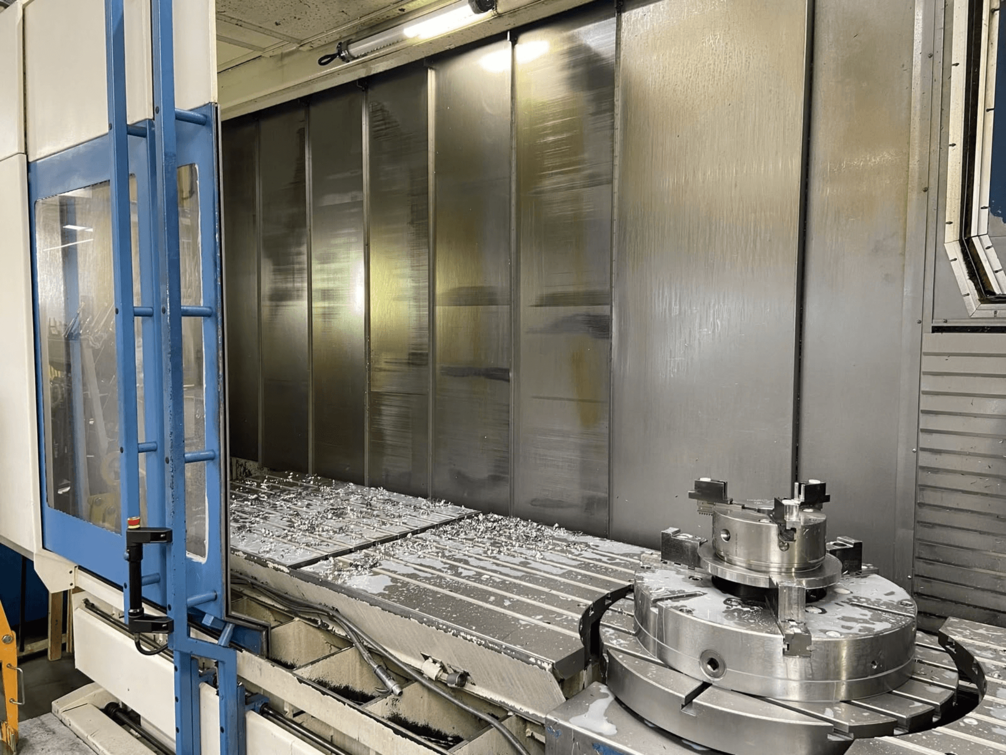 Vertikální obráběcí centrum AXA VHC-3-3000-M/E, pohled zepředu, na kterém jsou vidět frézované kovové třísky na pracovním stole a řezné nástroje.