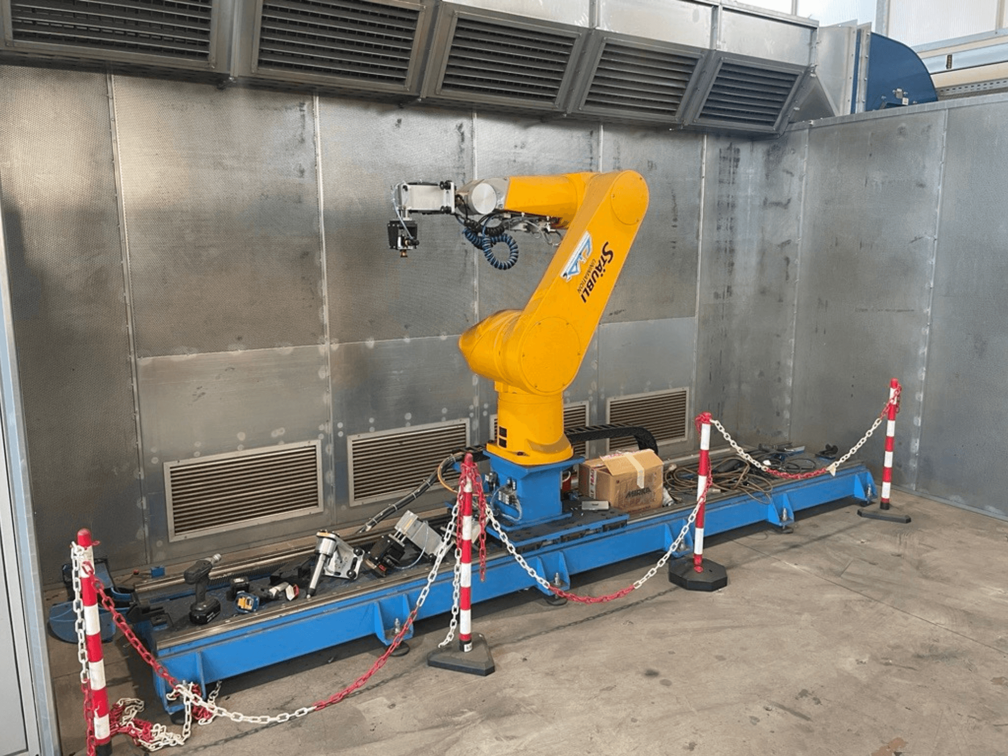 Robotické rameno STAUBLI CS8C-RX160 v dílně, boční pohled, s kabely a různými nástroji na modré základně obklopené ochrannými bariérami.
