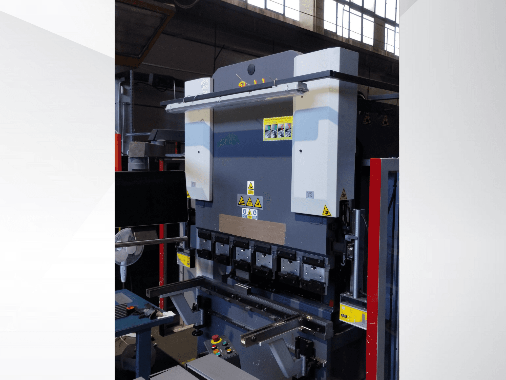 Lisovací stroj, pohled zepředu. Má viditelné ovládací prvky, bezpečnostní nálepky a několik držáků matric pro přesné ohýbání.