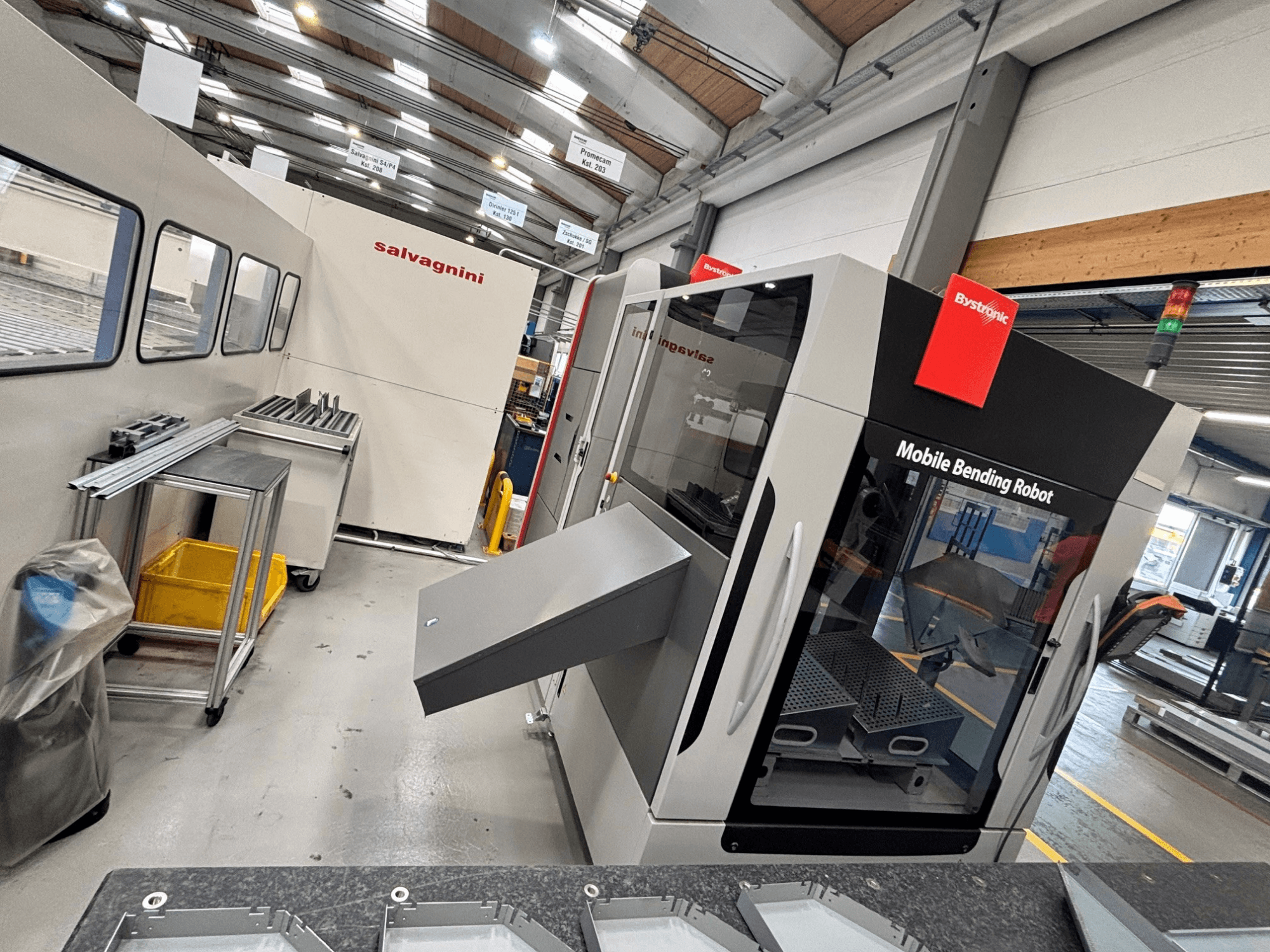 Ohýbačka Bystronic Xpert 40/1030, pohled zepředu znázorňující mobilního ohýbacího robota a pracovní prostor s kovovými díly na stole.