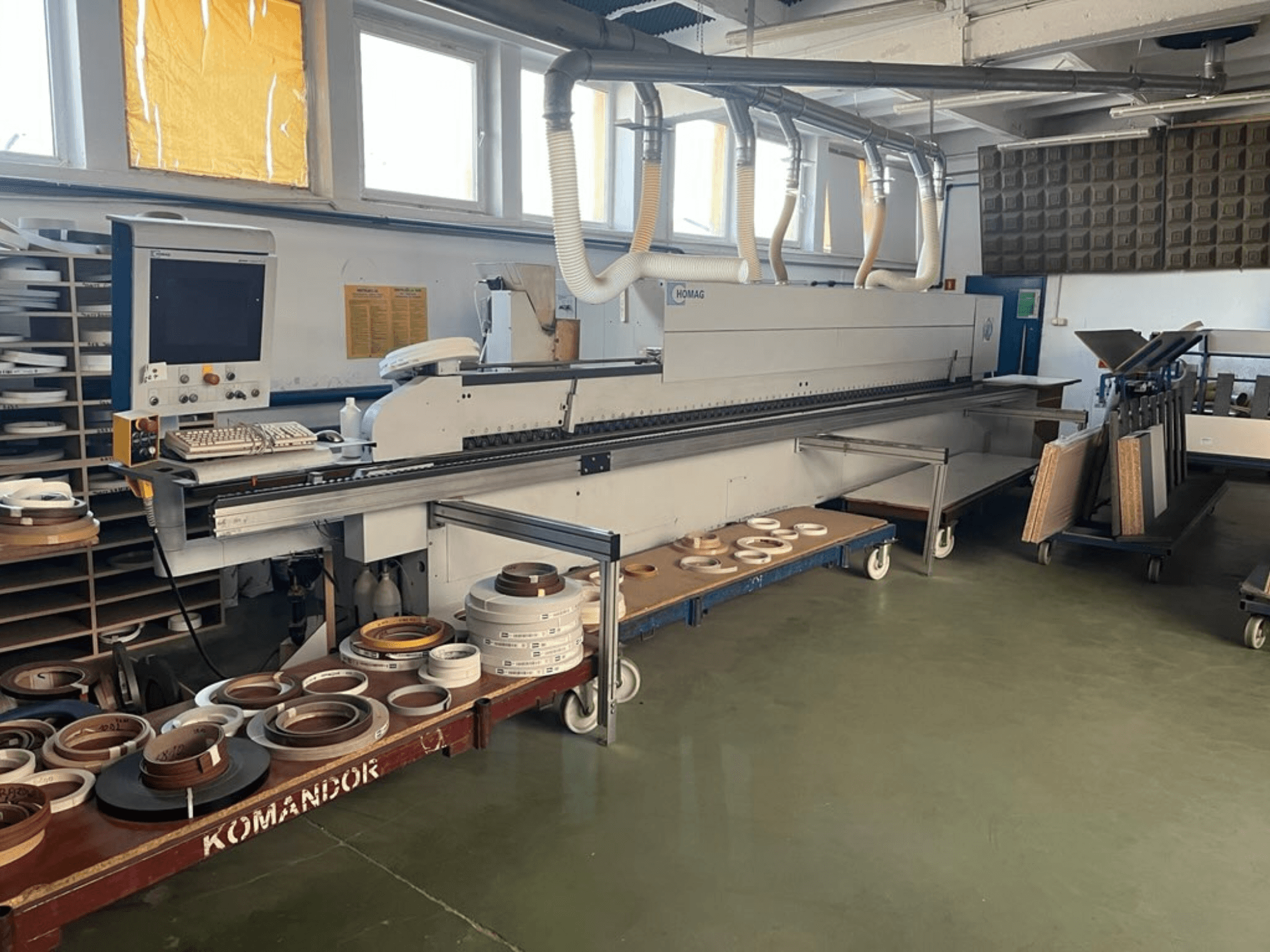 Olepovačka hran HOMAG v dílně s ovládacími prvky a různými olepovacími materiály na modrém vozíku. Boční pohled.