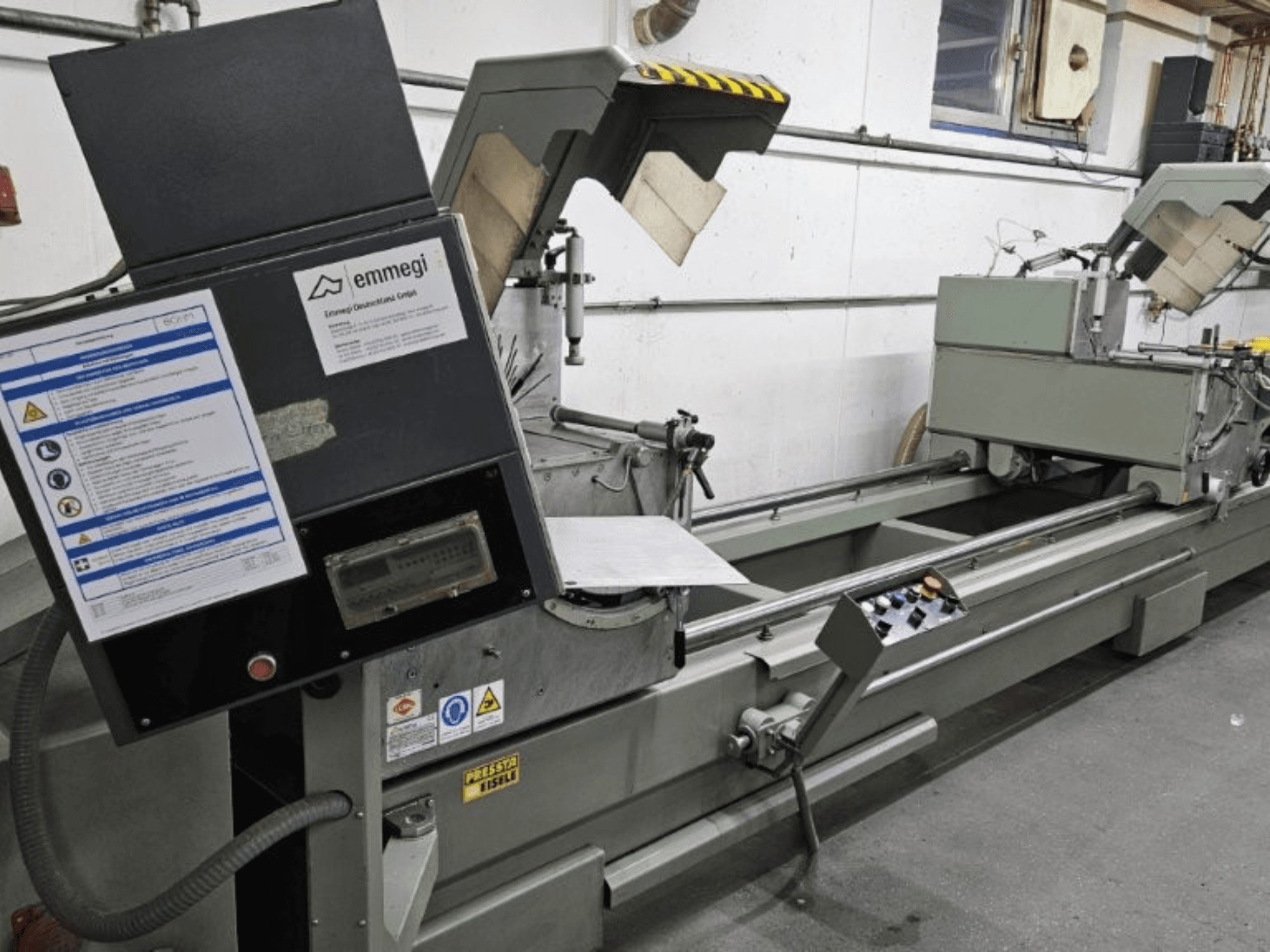 Průmyslový řezací stroj Emmegi, pohled zepředu, s ovládacím panelem, bezpečnostními štítky a provozními prvky.