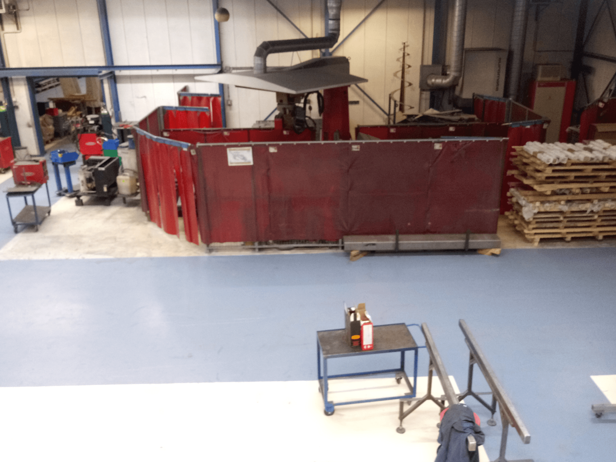 Průmyslové pracoviště s červenými svářecími závěsy, stroji a paletami; pohled shora ukazuje uspořádání a vybavení.