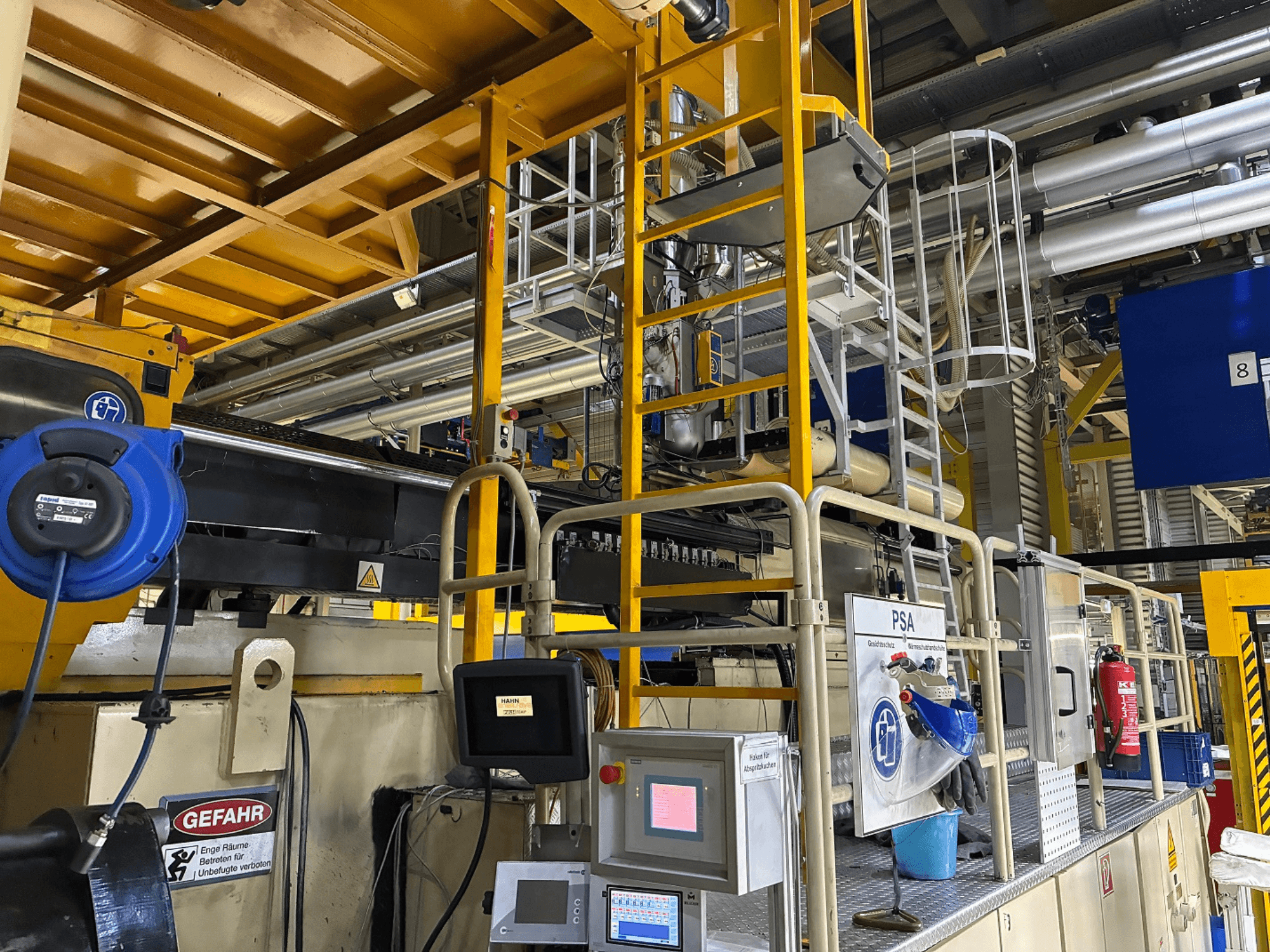 Ovládací panel průmyslového stroje s modrým bezpečnostním vybavením a žlutým lešením, boční pohled zobrazující potrubí a výstražné značky.