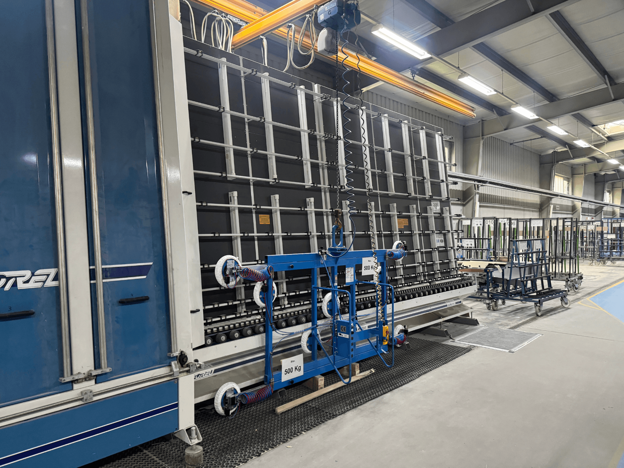 Průmyslové zařízení společnosti Drel s velkým rámem a zvedacím mechanismem, zobrazené z bočního pohledu v továrním prostředí.