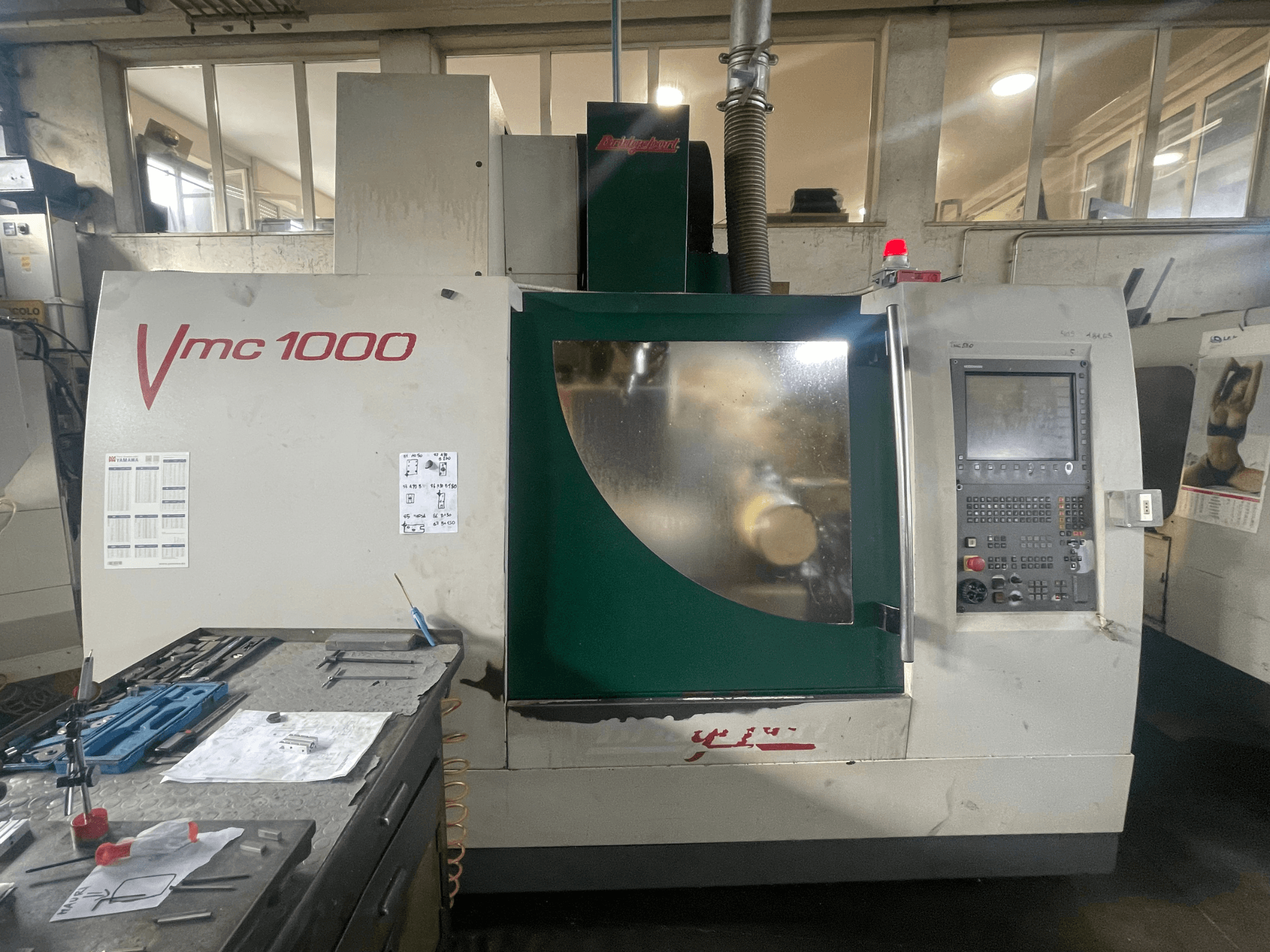 Vertikální obráběcí centrum Bridgeport VMC 1000/30, pohled zepředu, na kterém je vidět ovládací panel a hlavní těleso stroje, s nástroji v blízkosti.