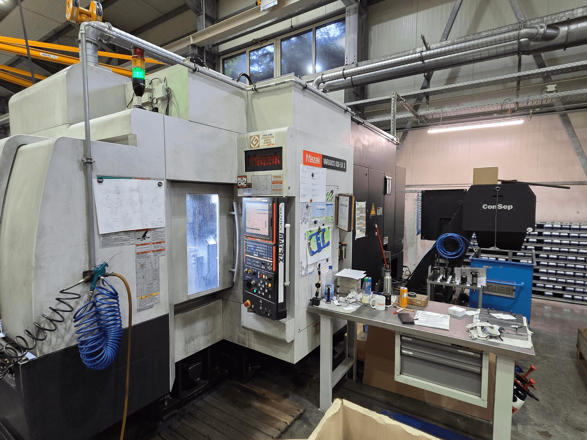 CNC stroj Mazak Variaxis 630-5X v dílně, čelní pohled s ovládacím panelem, hadicí a příslušenstvím na bočním stole.