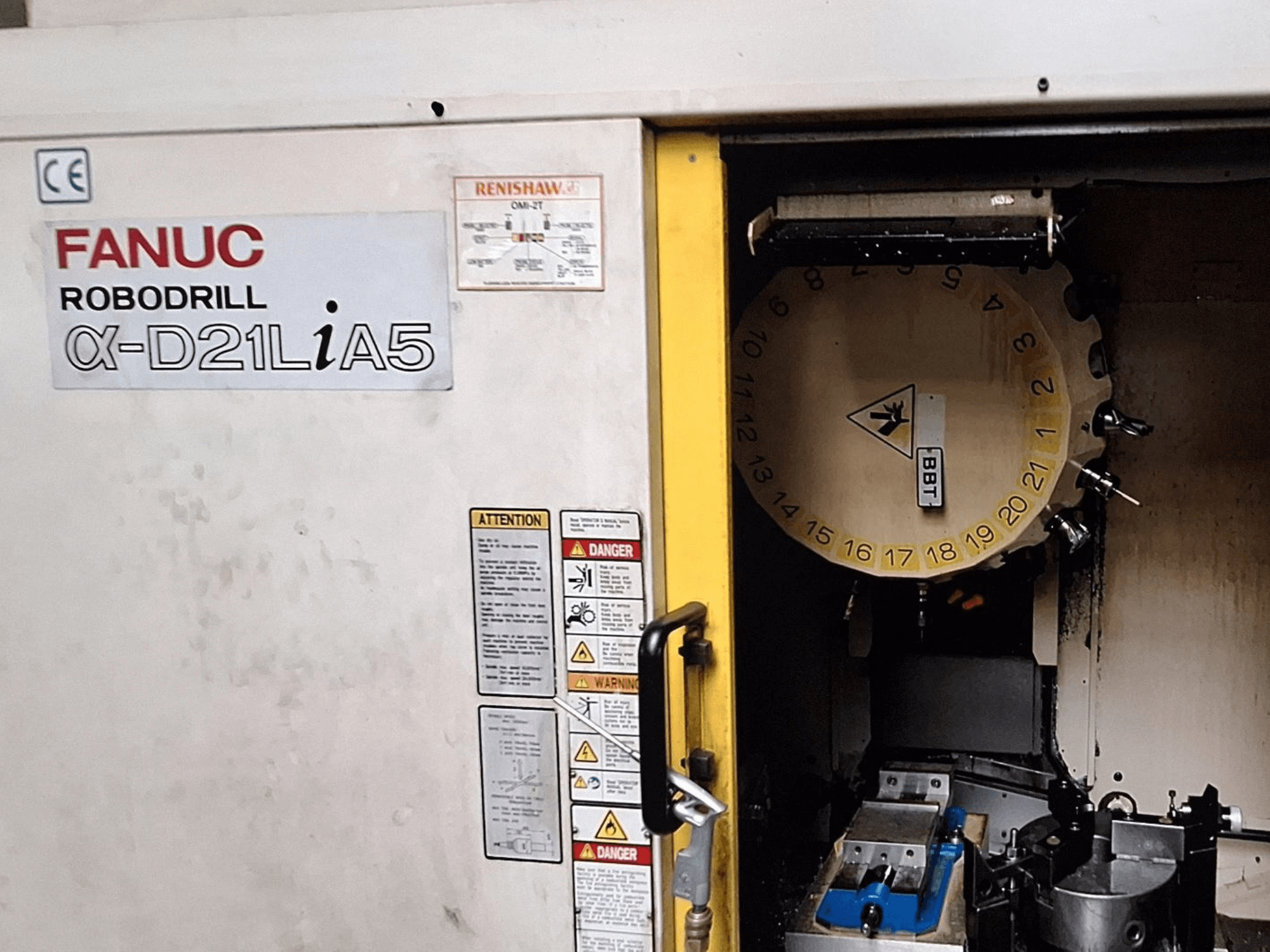 Robovrták FANUC α-D21LiA5, pohled zepředu. Zobrazuje karusel nástrojů a obráběcí prostor se svorkami a upozorněními na dveřích.