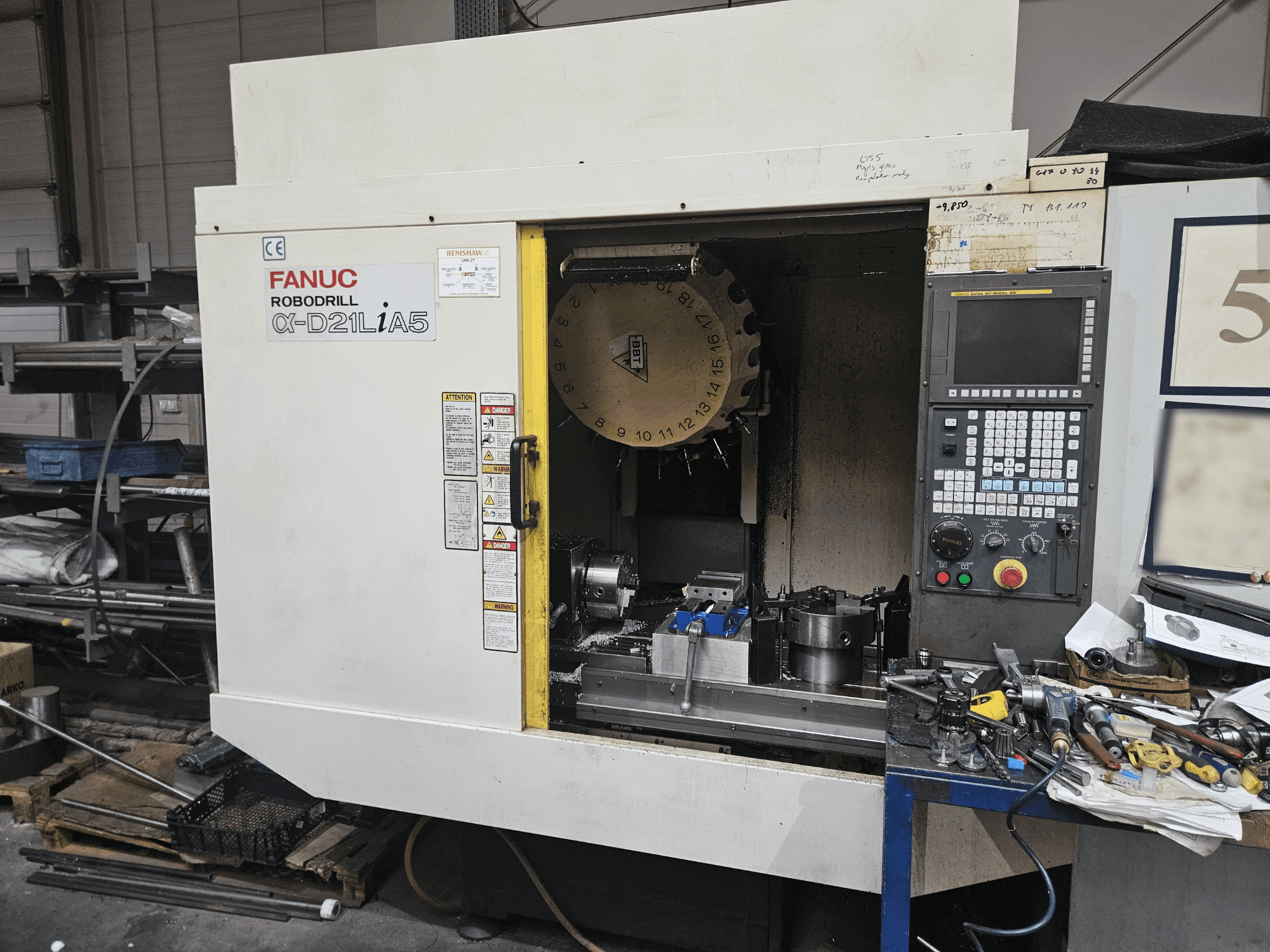 Robovrták FANUC α-D21LiA5, pohled zepředu na držák nástrojů a ovládací panel, obklopený nástroji a zařízením v dílně.