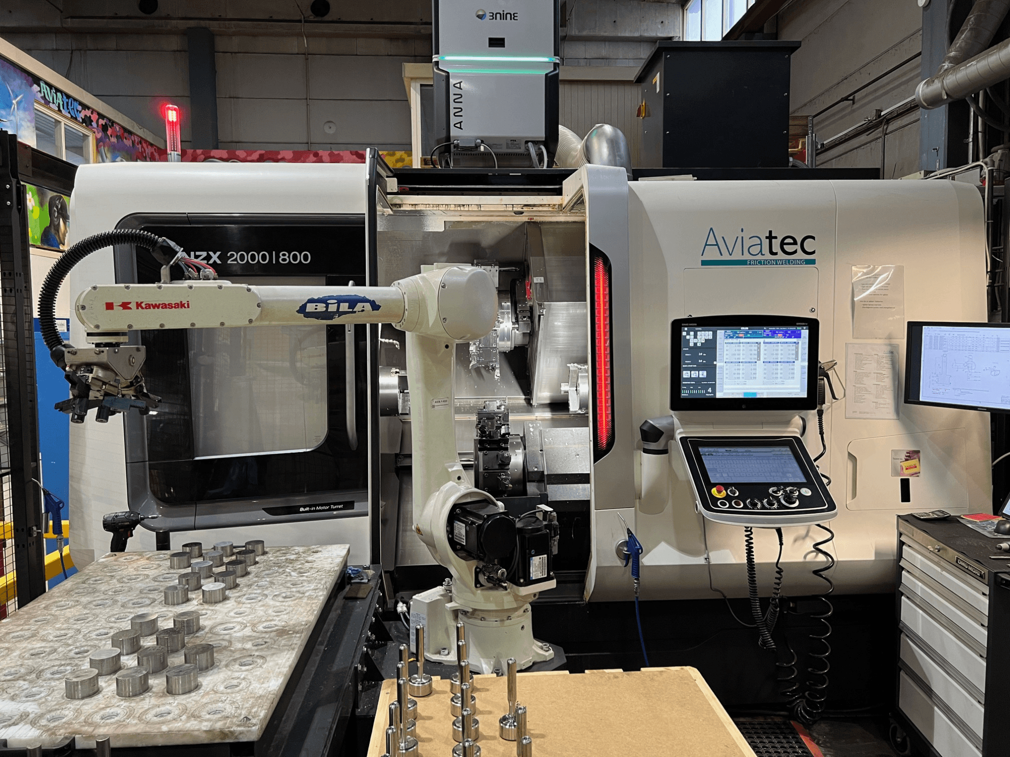 Horizontální soustruh DMG MORI NZX 2000/800SY s robotickým ramenem Kawasaki, ovládacím panelem a kovovými součástmi na pracovním stole.