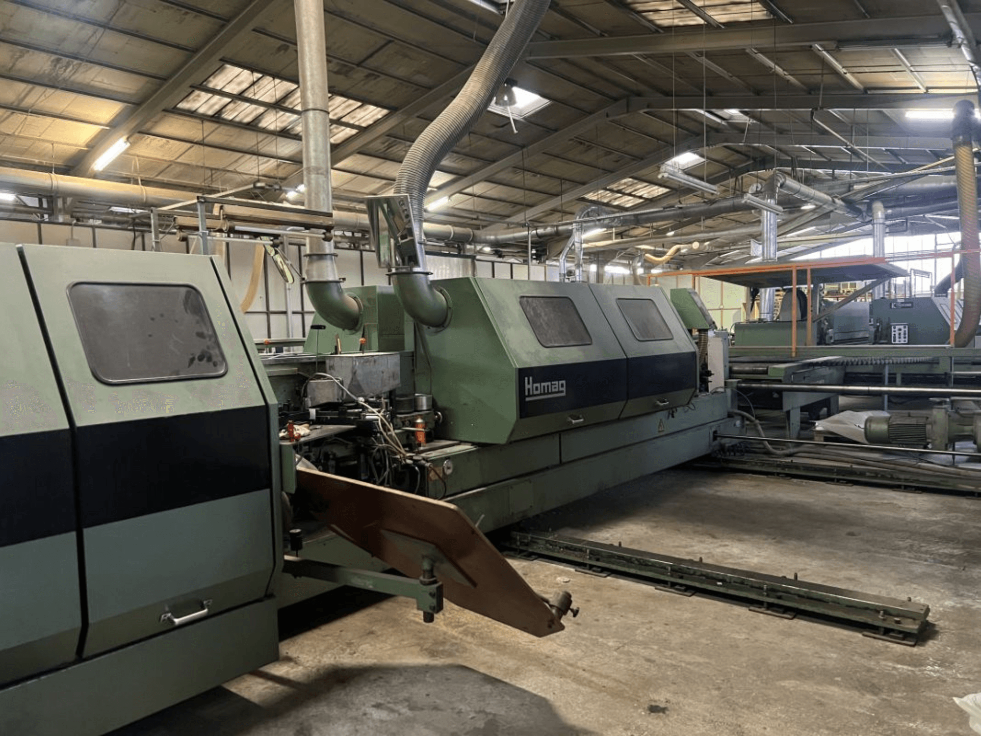 Průmyslový stroj Homag v zelené barvě, boční pohled, s viditelnými provozními prvky a ovládacím panelem ve skladu.