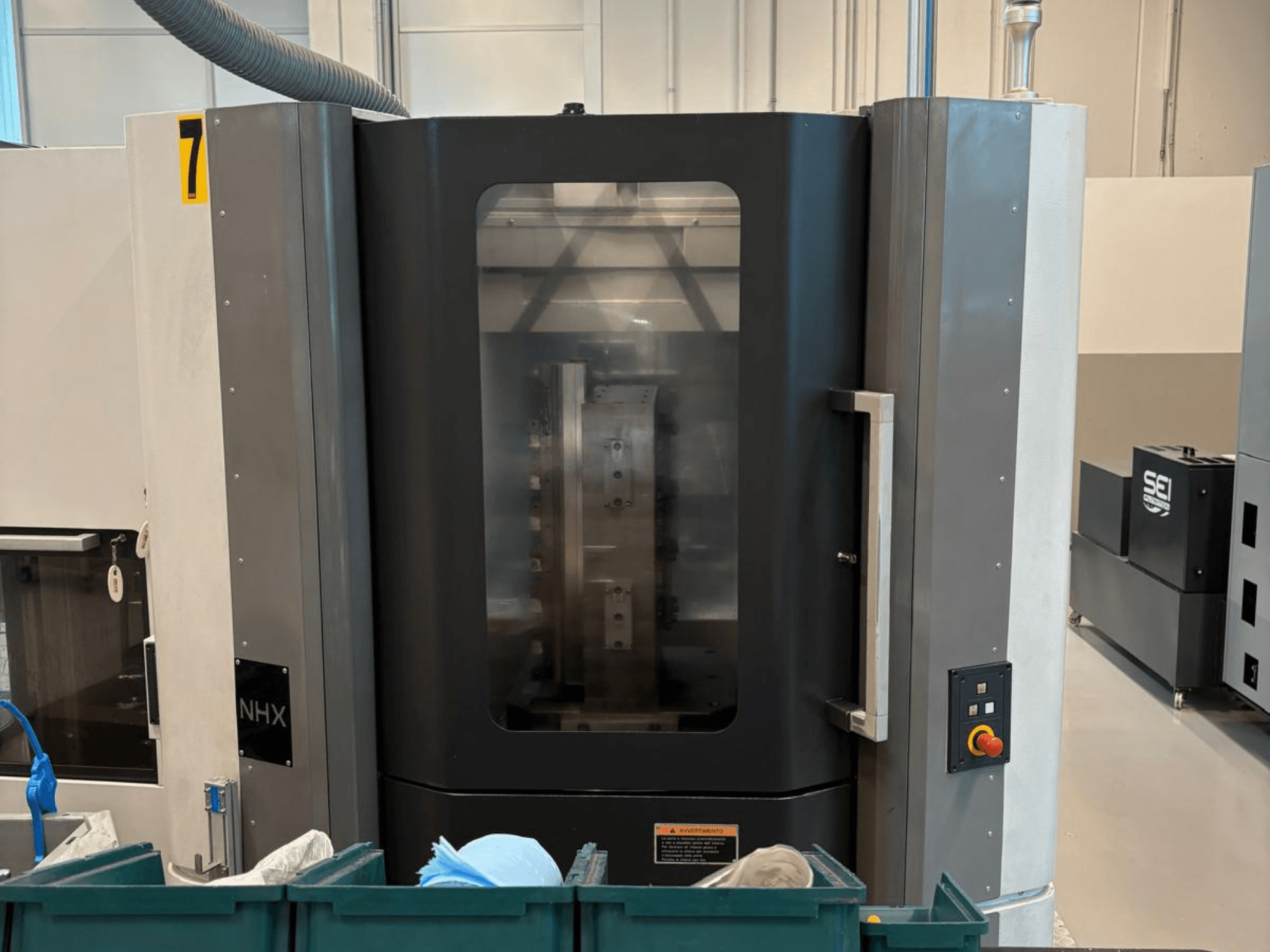 Stroj NHX CNC v čelním pohledu, zobrazující ovládací panel a přehledný displej; umístěný v dílně se zelenými zásobníky v blízkosti.