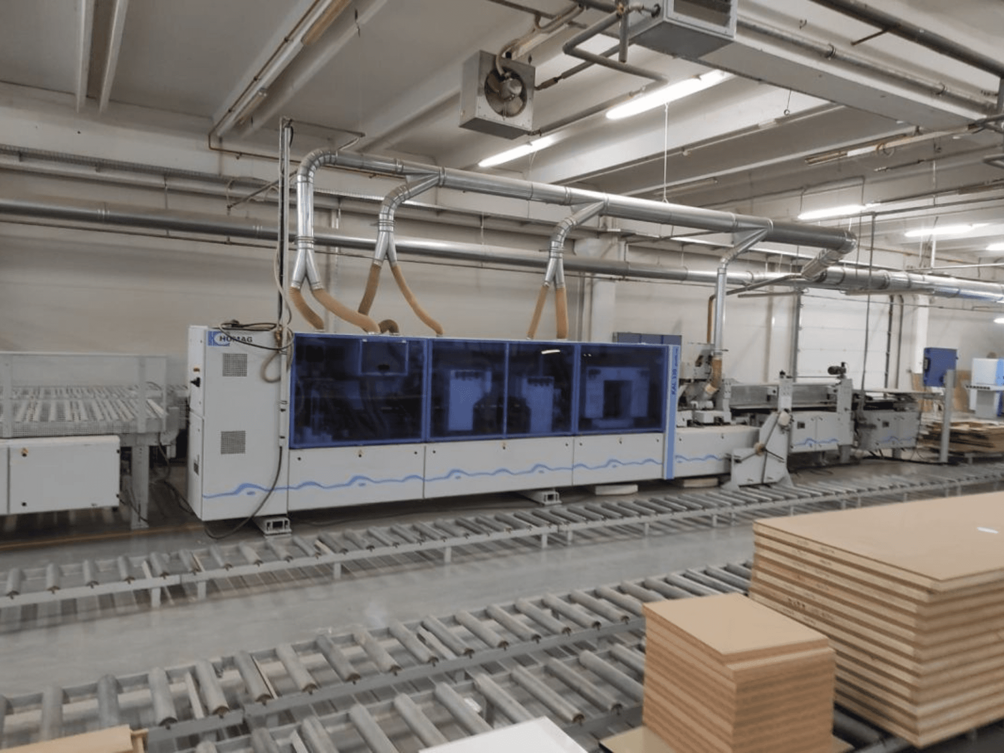 Výrobní stroj HOMAG v továrně, pohled zepředu, s modrým pláštěm a dopravníkovým systémem s naskládanými překližkovými deskami.