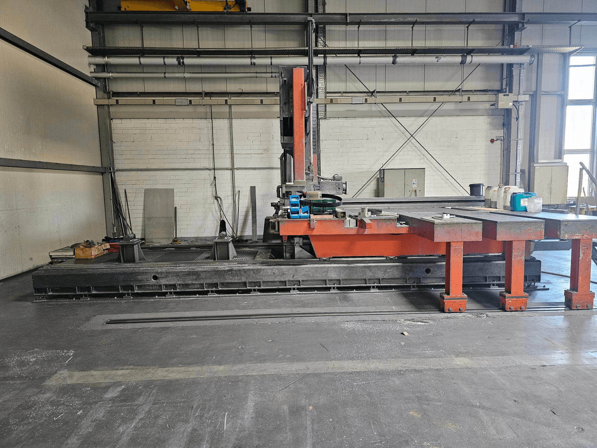 Průmyslový řezací stroj v otevřené dílně, oranžový a černý s velkým stolem; boční pohled ukazuje příslušenství a pracovní prostor.