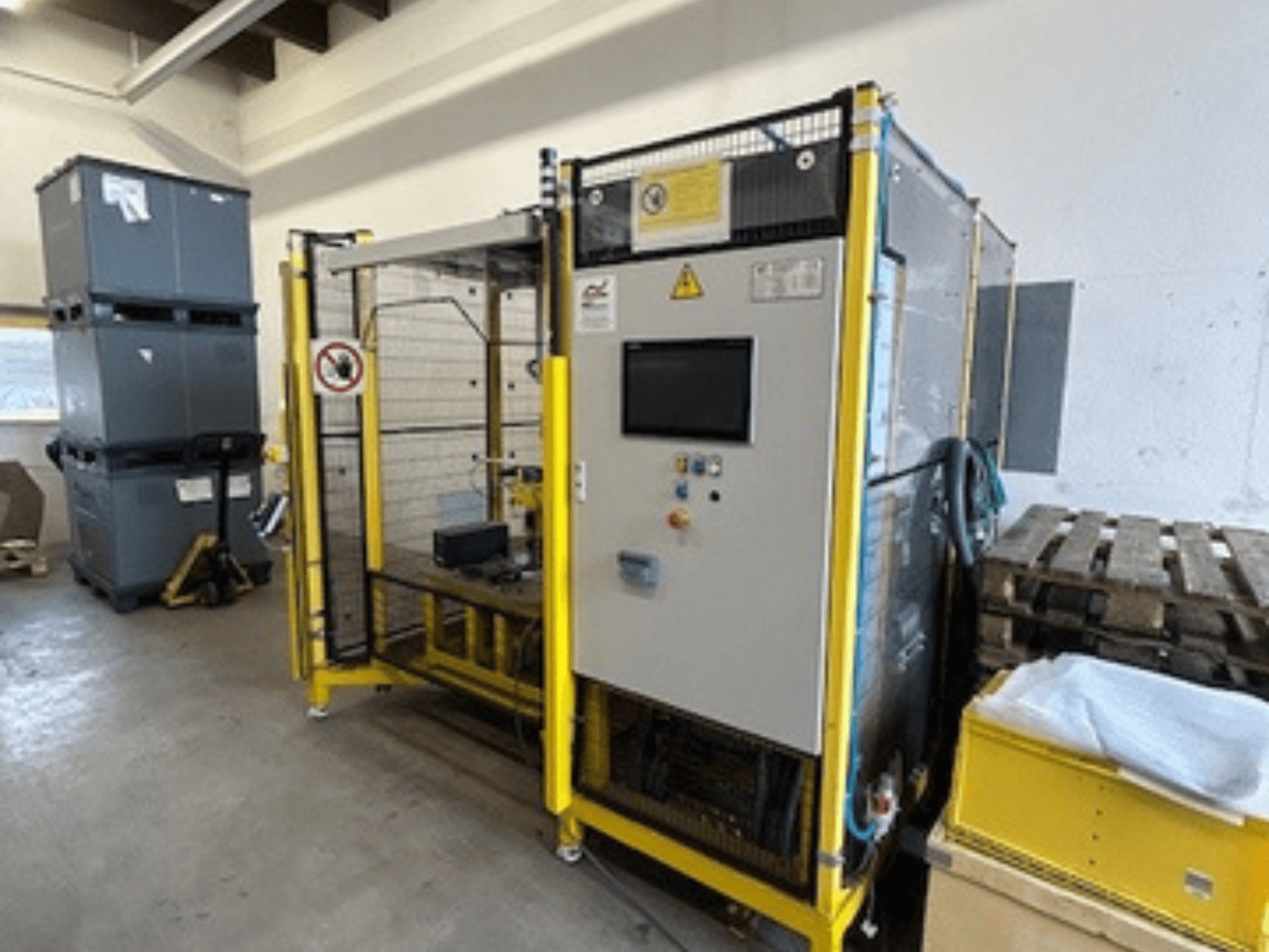 Průmyslový stroj s ovládacím panelem a bezpečnostními prvky, pohled zepředu; žlutý rám, šedé zásobníky a palety v pozadí.