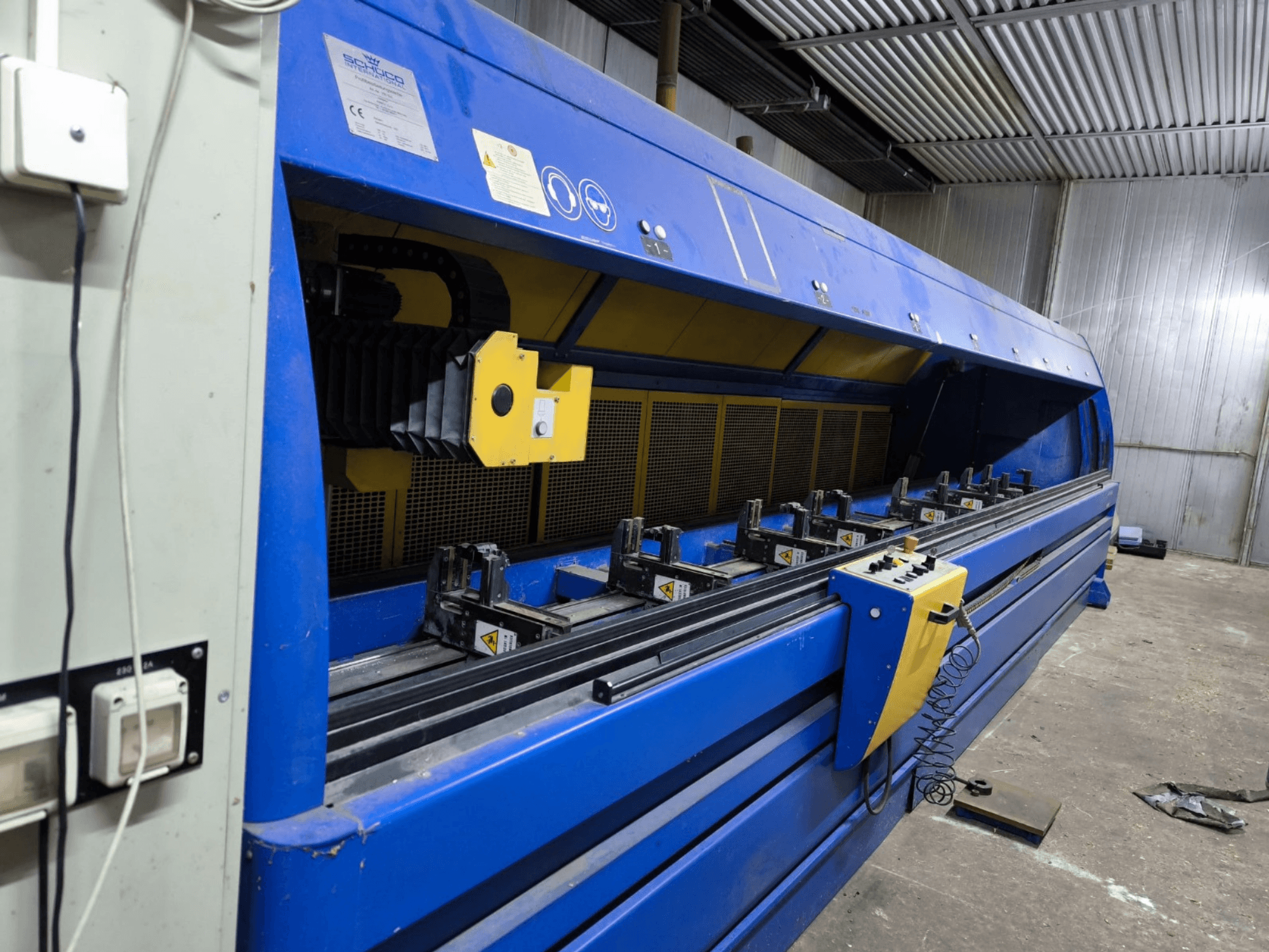 Průmyslový řezací stroj Schneider v modré barvě, pohled z boku, s viditelným ovládacím panelem, pásy a několika upínacími stanicemi.