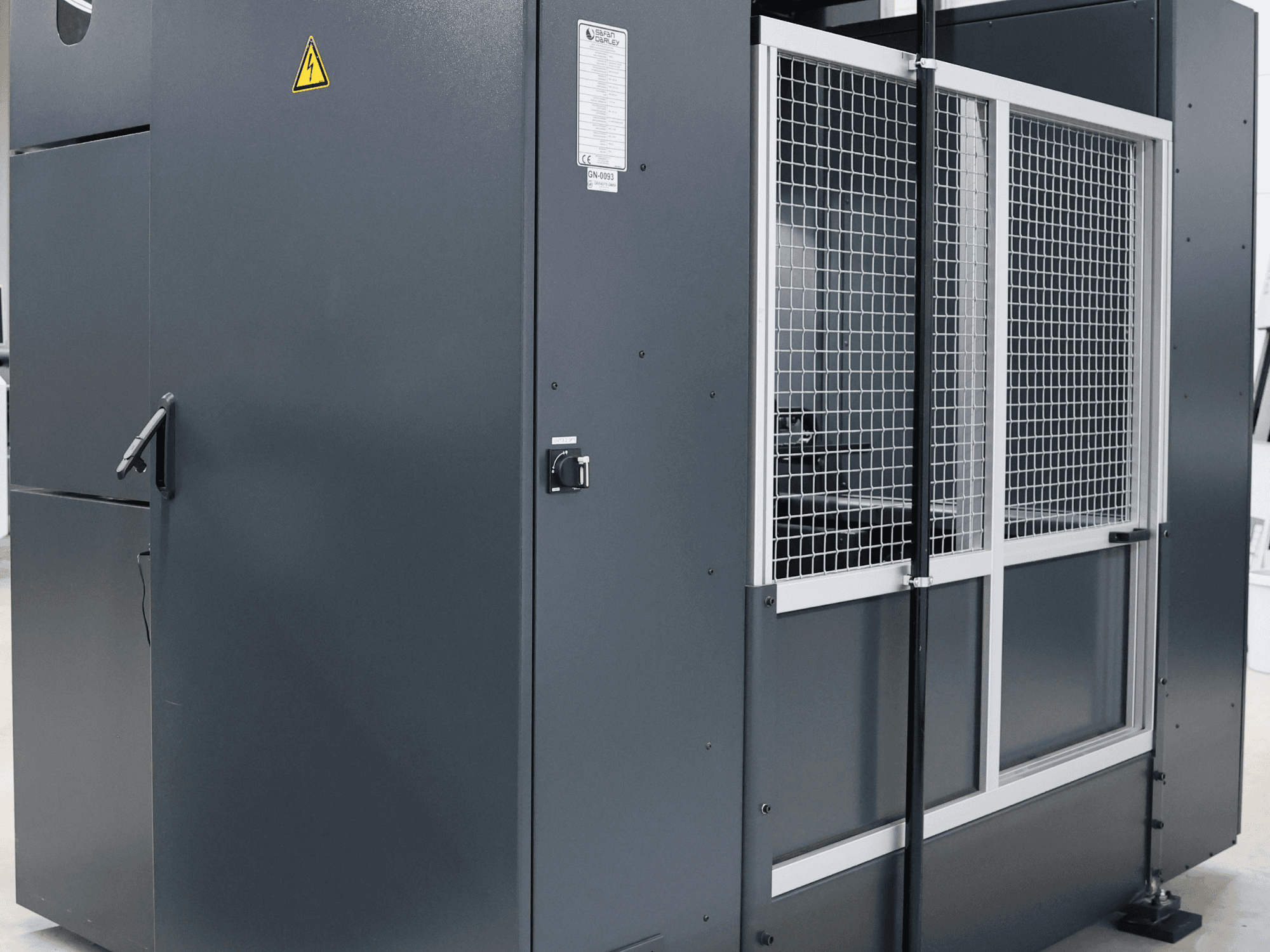 Šedý průmyslový stroj s bezpečnostním upozorněním, pohled zepředu ukazuje síťová dvířka a ovládací panel, model označen.