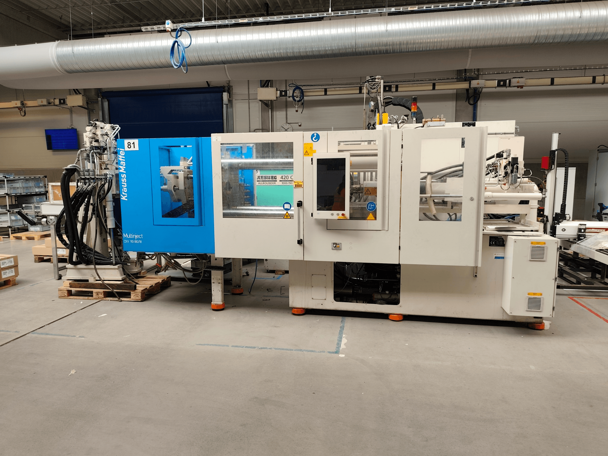 Hydraulický vstřikovací stroj Krauss Maffei 110-180-180 CX V, boční pohled, na kterém je vidět modrobílé tělo stroje, ovládací panel a konektory.