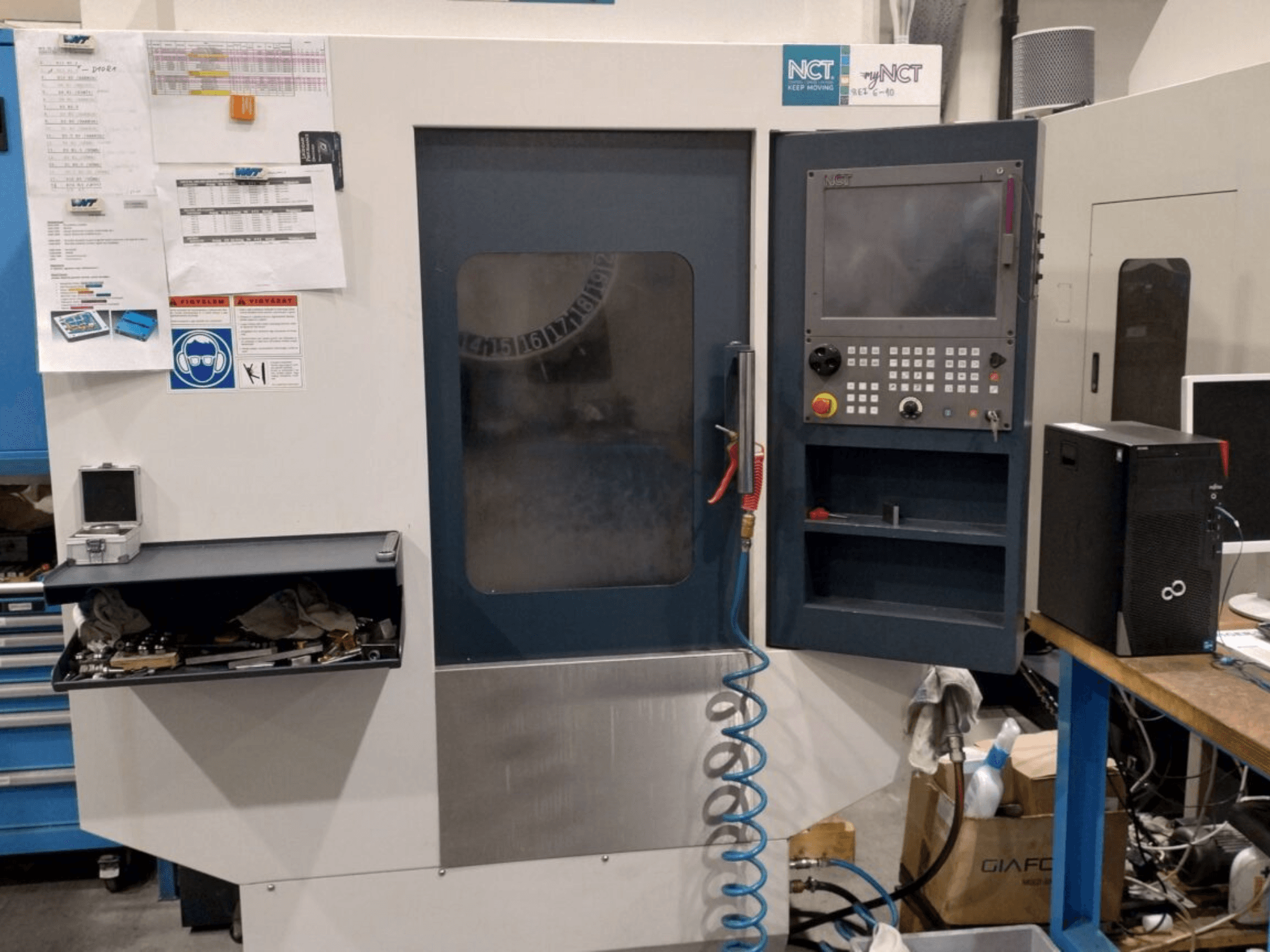 Stroj NCT CNC, pohled zepředu, ovládací panel s numerickou klávesnicí, nástroje v úložné zásuvce a návod k obsluze na stěně.