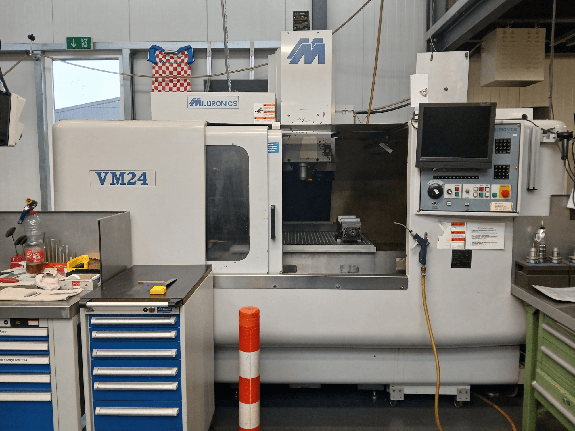 CNC frézka Milltronics VM24 zobrazená zepředu, s ovládacím panelem, úložným prostorem pro nástroje a nastavením pracovní plochy.