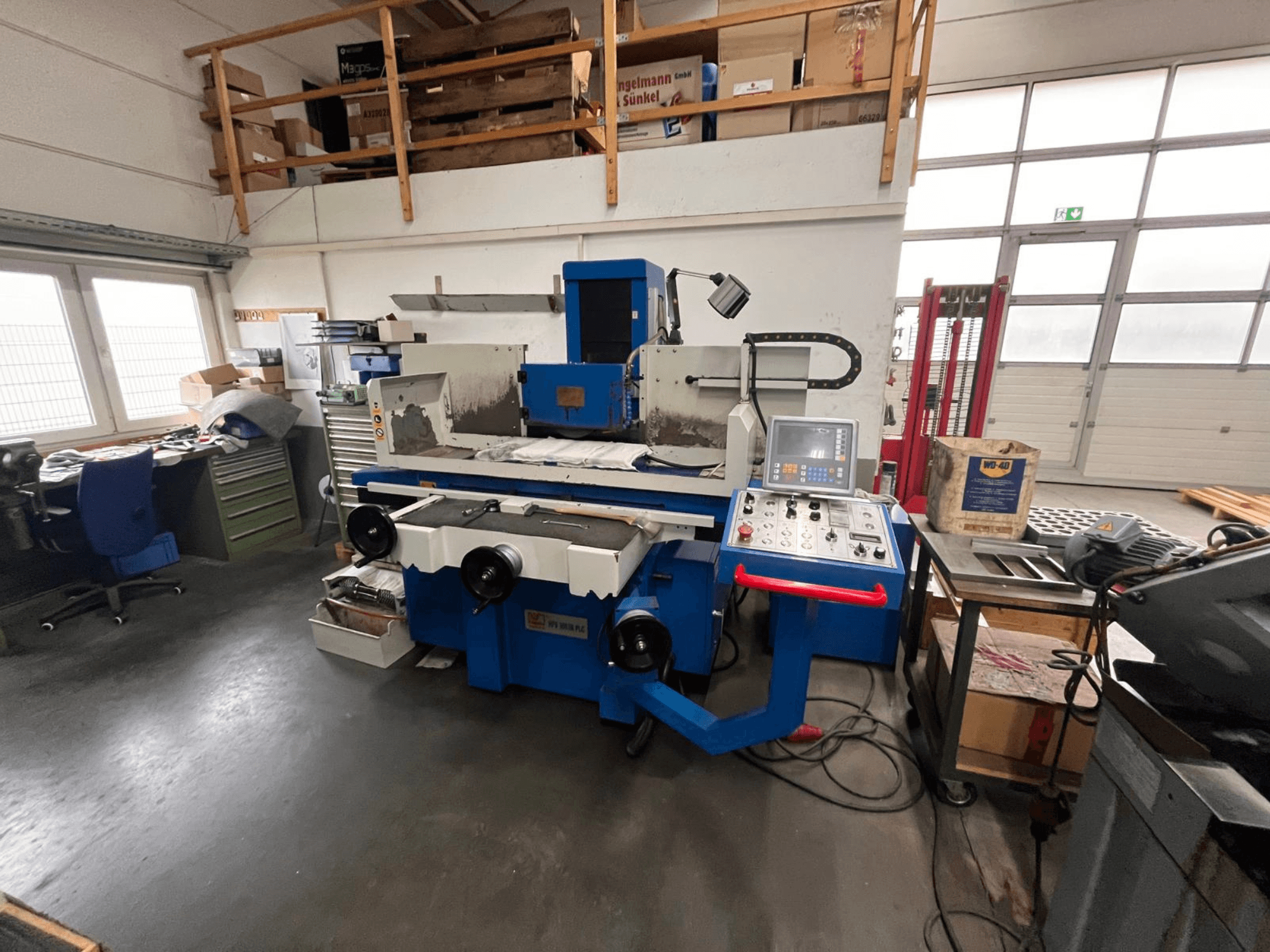 Modrá bruska na plocho s digitálním ovládacím panelem, pohled zepředu, zobrazující nepořádek na pracovišti a zelenou skříňku na nářadí.