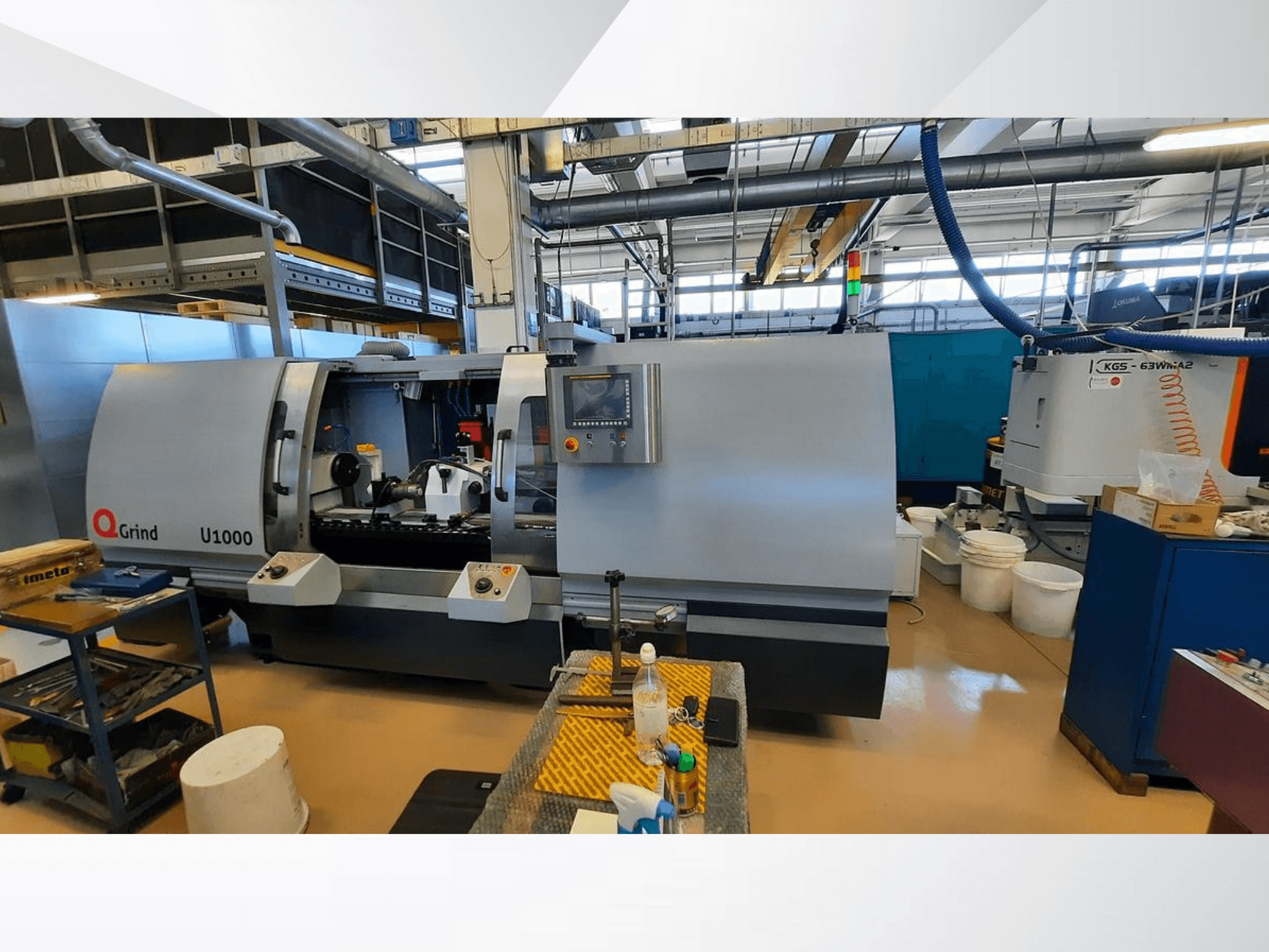 Stroj Q-Grind U1000 v dílně, pohled zepředu s ovládacím panelem a otevřeným řezacím prostorem, obklopený zařízením.