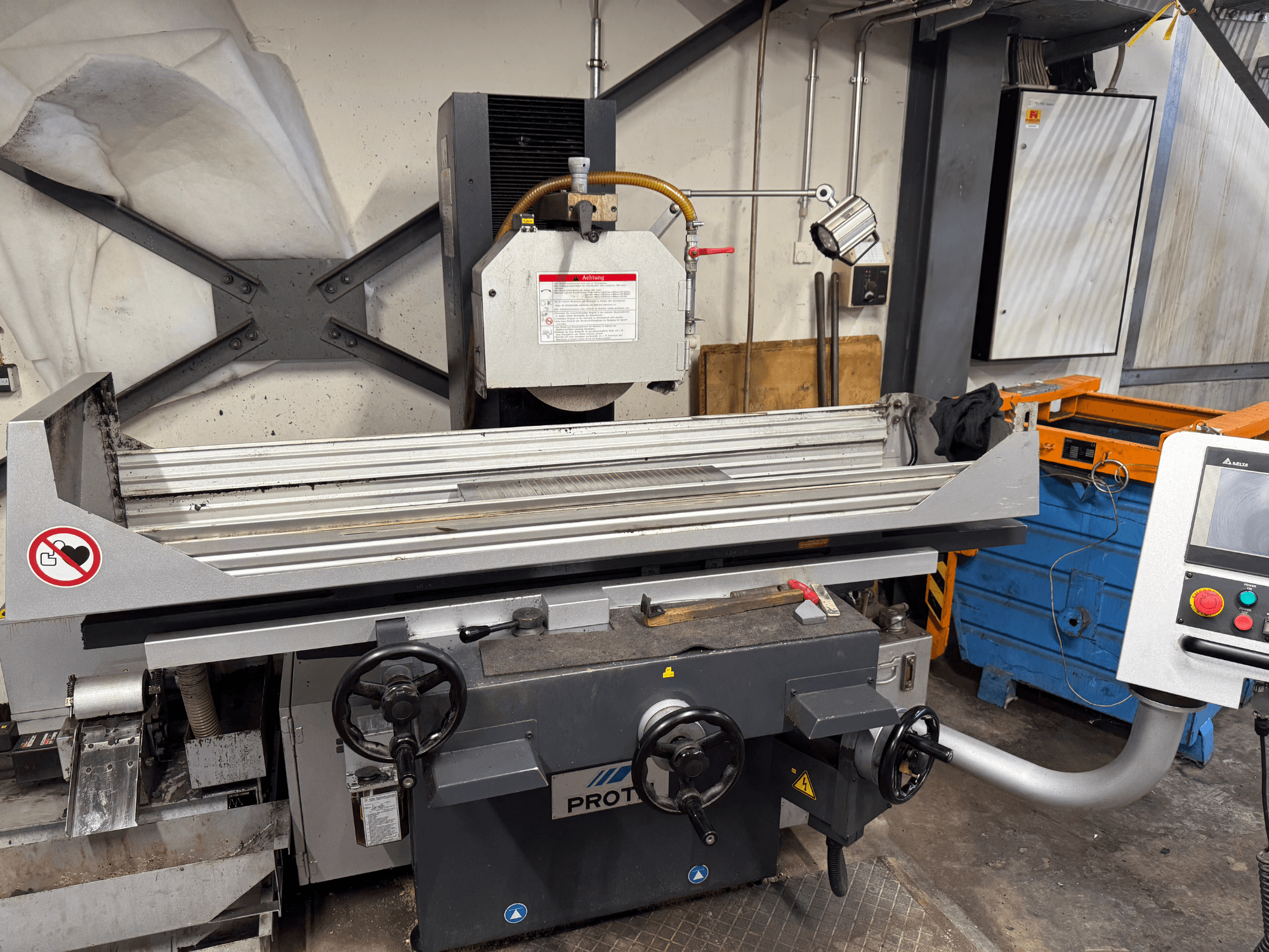 Stroj PROTEC, pohled zepředu, zobrazující brusku s ručním ovládáním, bezpečnostní upozornění a chladicí systém.