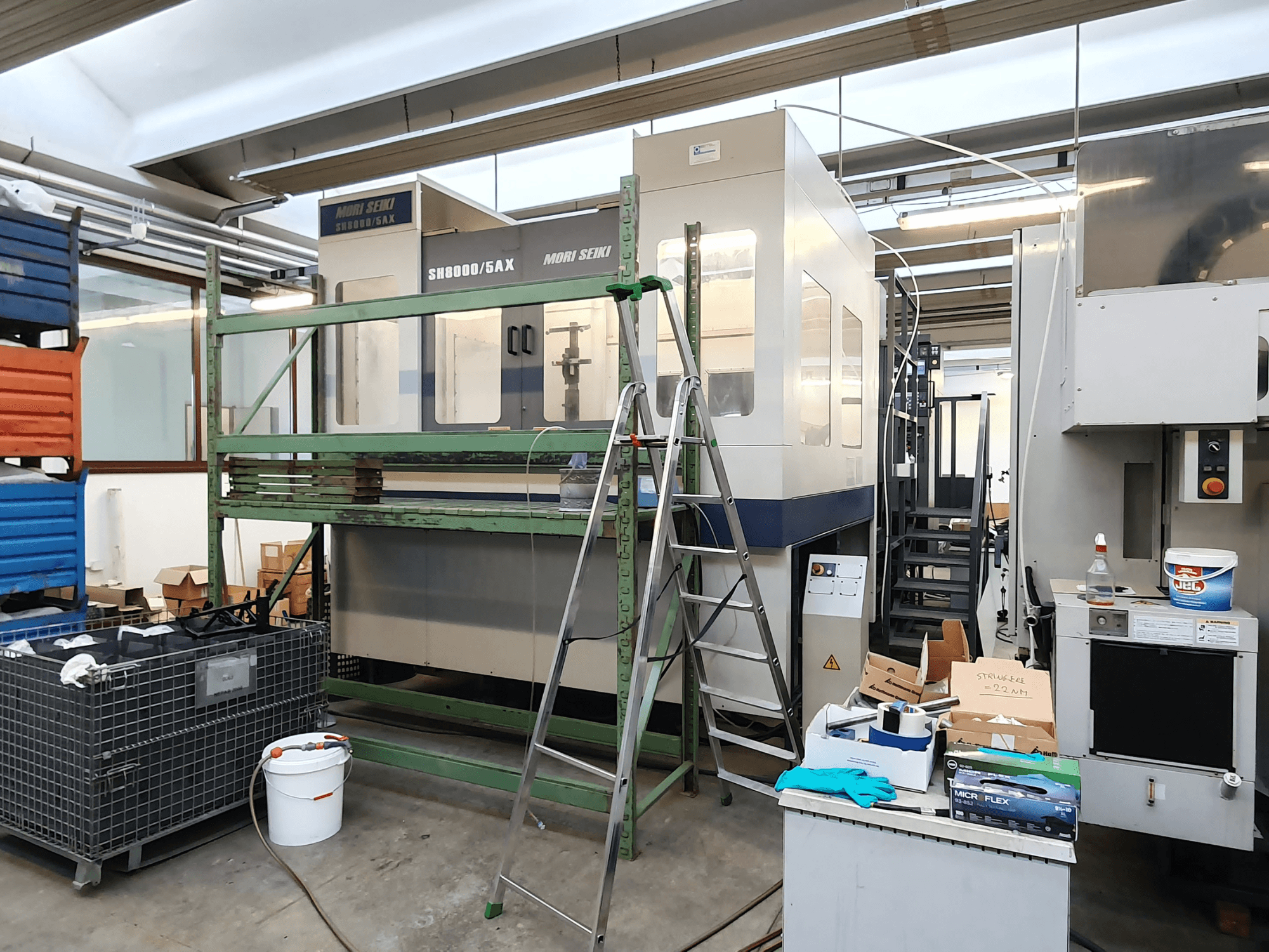 CNC stroj Mori Seiki SH-8000/5AX v dílně, pohled zepředu, s dílenským nářadím a úložnými boxy poblíž.