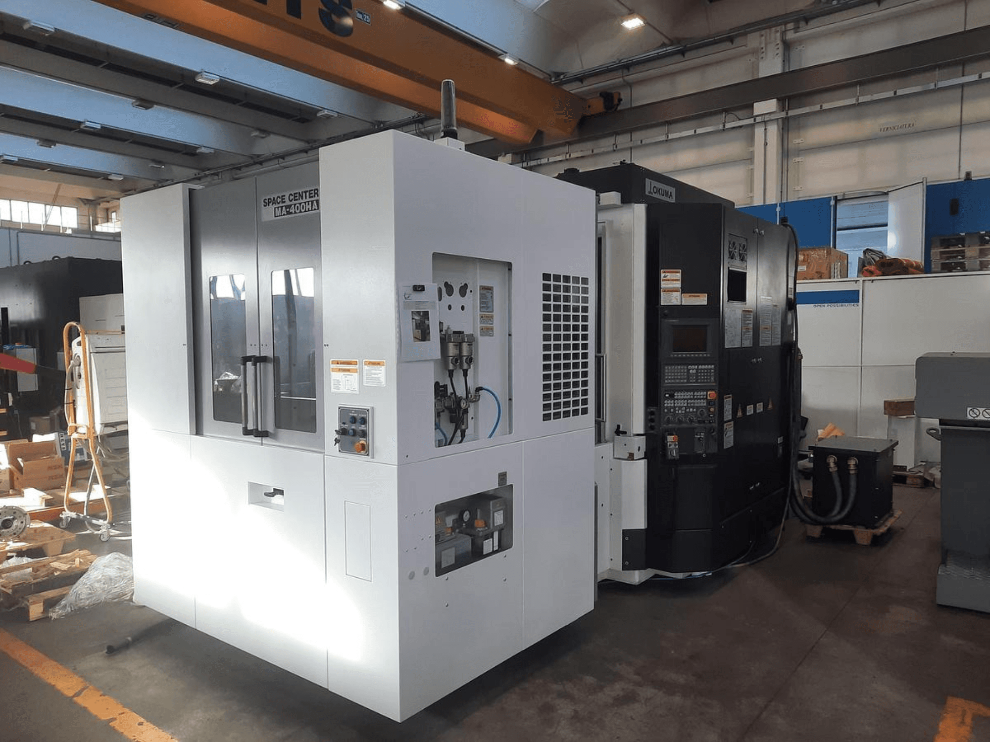 Pohled zepředu na stroje SPACE CENTER IM-400HAA a OKUMA CNC s různými ovládacími prvky a štítky výrobce.
