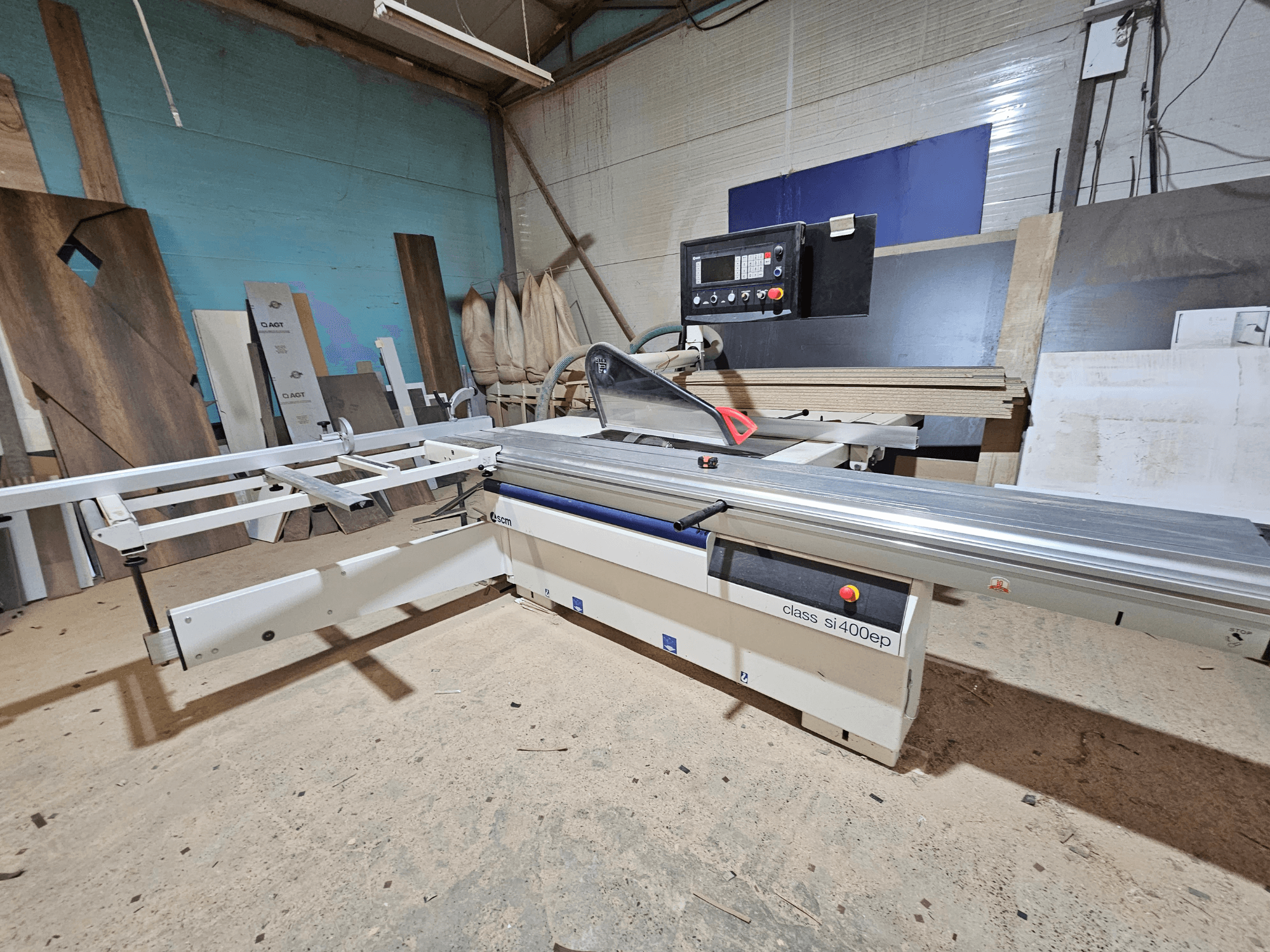 Desková pila třídy SI400EP, pohled zepředu, s nastavitelným plotem, postavená na pískové podlaze dílny, obklopená dřevěnými deskami.