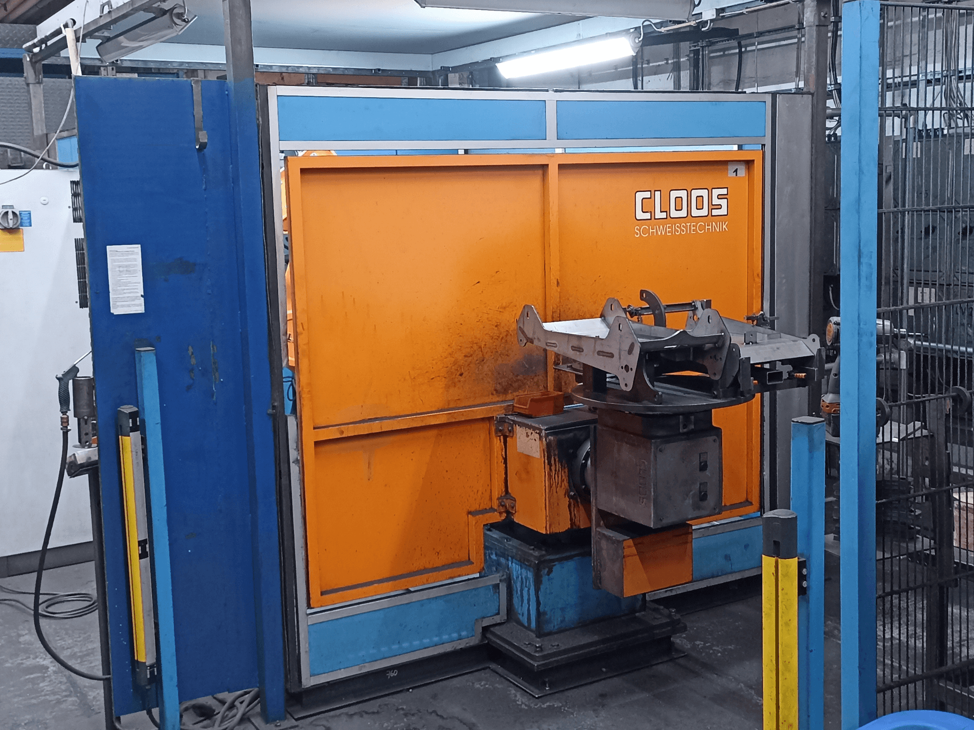 Svařovací robot CLOOS Romat 320 v dílně, pohled zepředu, s oranžovým ovládacím panelem a kovovým obrobkem na otočné základně.