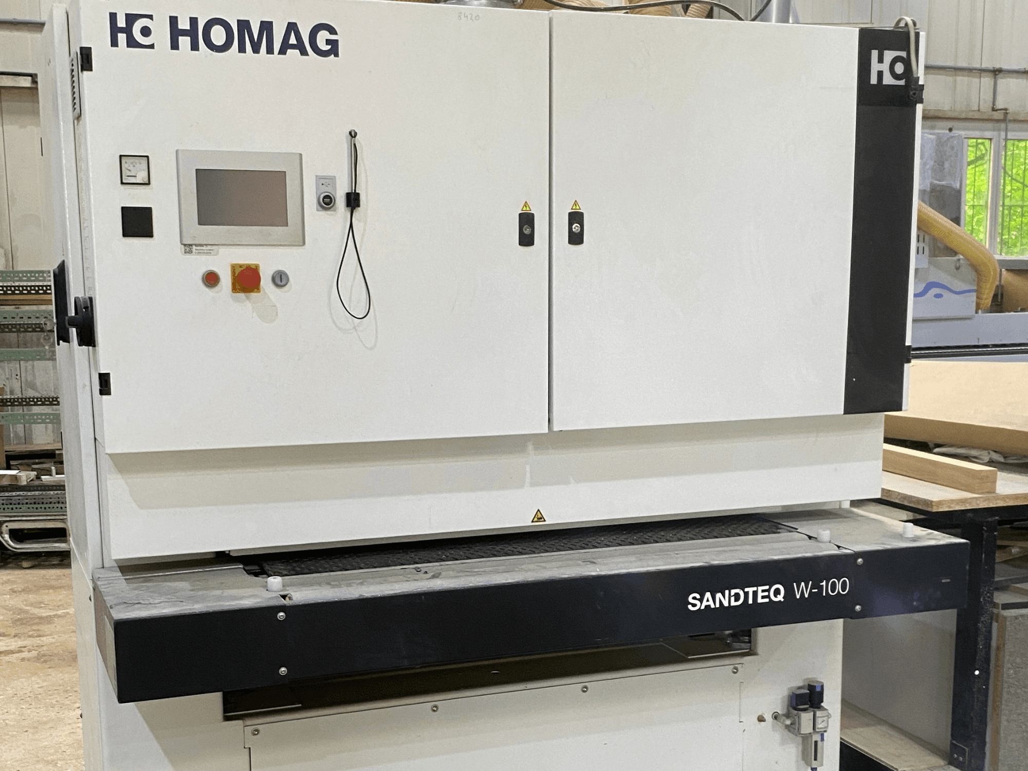 Pásová bruska HOMAG SANDTEQ W-100 v dílně; na snímku je vidět ovládací panel, přípojka pro odsávání prachu a oblast s brusným pásem. Pohled zepředu.