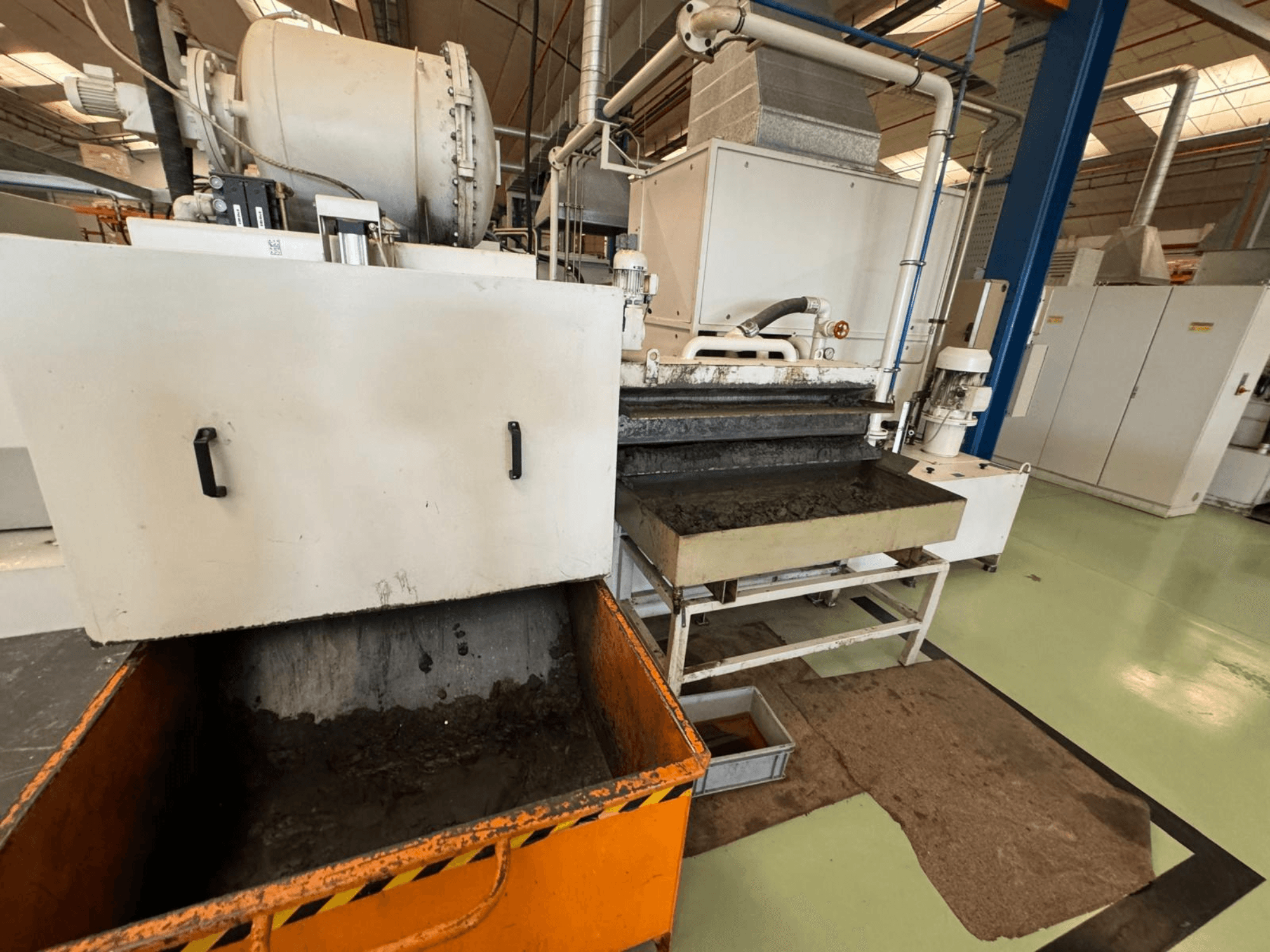 Průmyslové zpracovatelské zařízení s oranžovou nádobou na odpad, pohled zepředu; obsahuje motor, dopravníky a prostor pro údržbu.