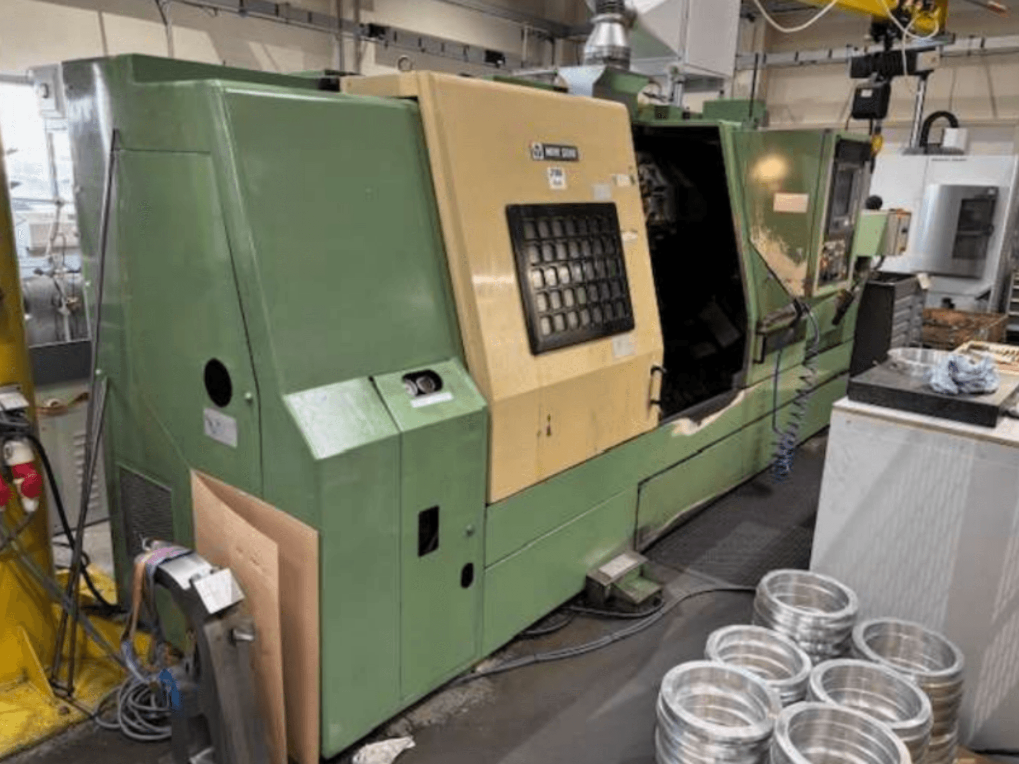 Zelený CNC soustruh Mazak, pohled zepředu, s ovládacím panelem a obráběnými hliníkovými díly na pracovním stole.