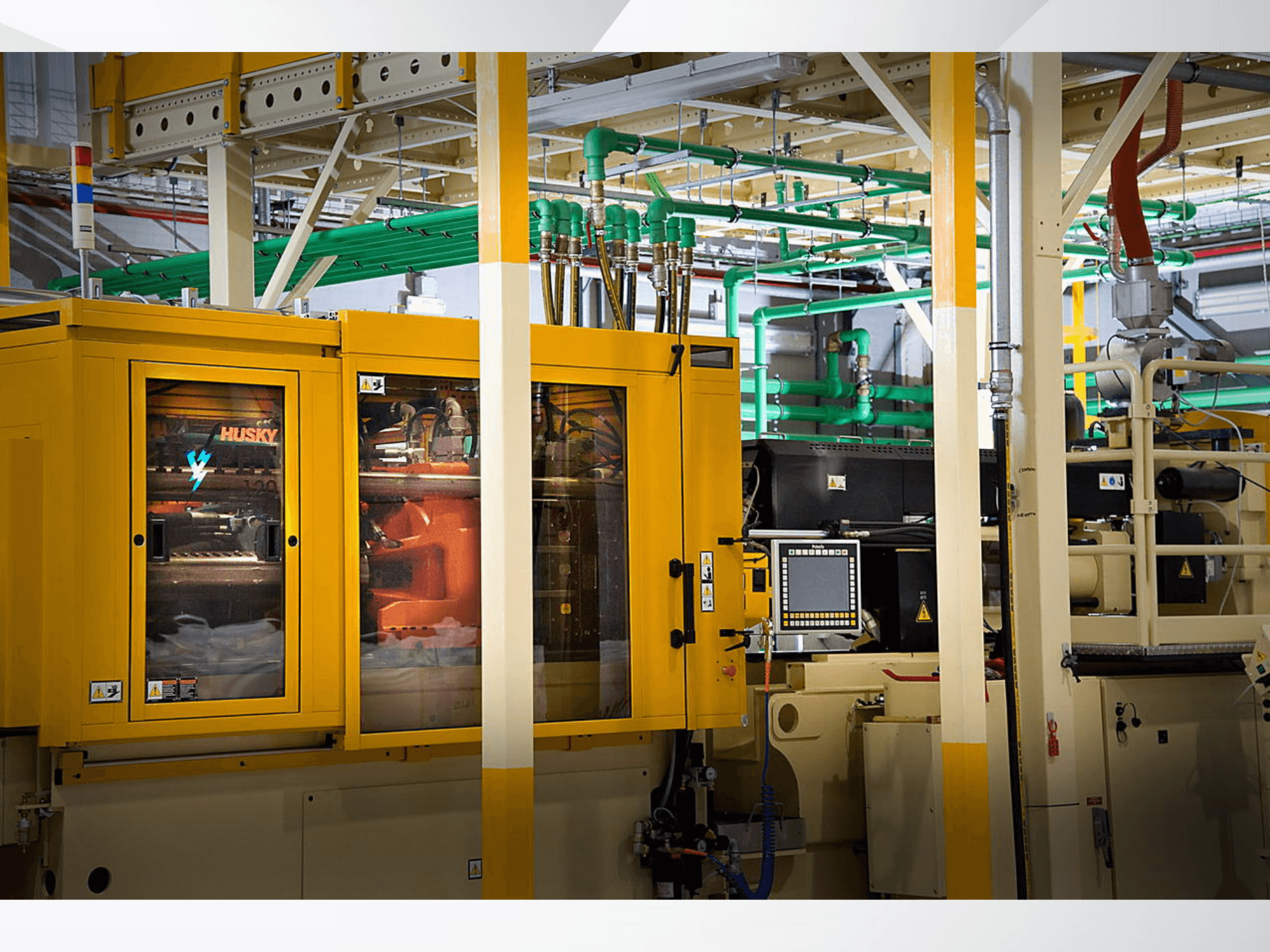 Průmyslový stroj Husky ve žluté barvě, čelní pohled na ovládací panel a vnitřní mechanismy na pozadí továrny.