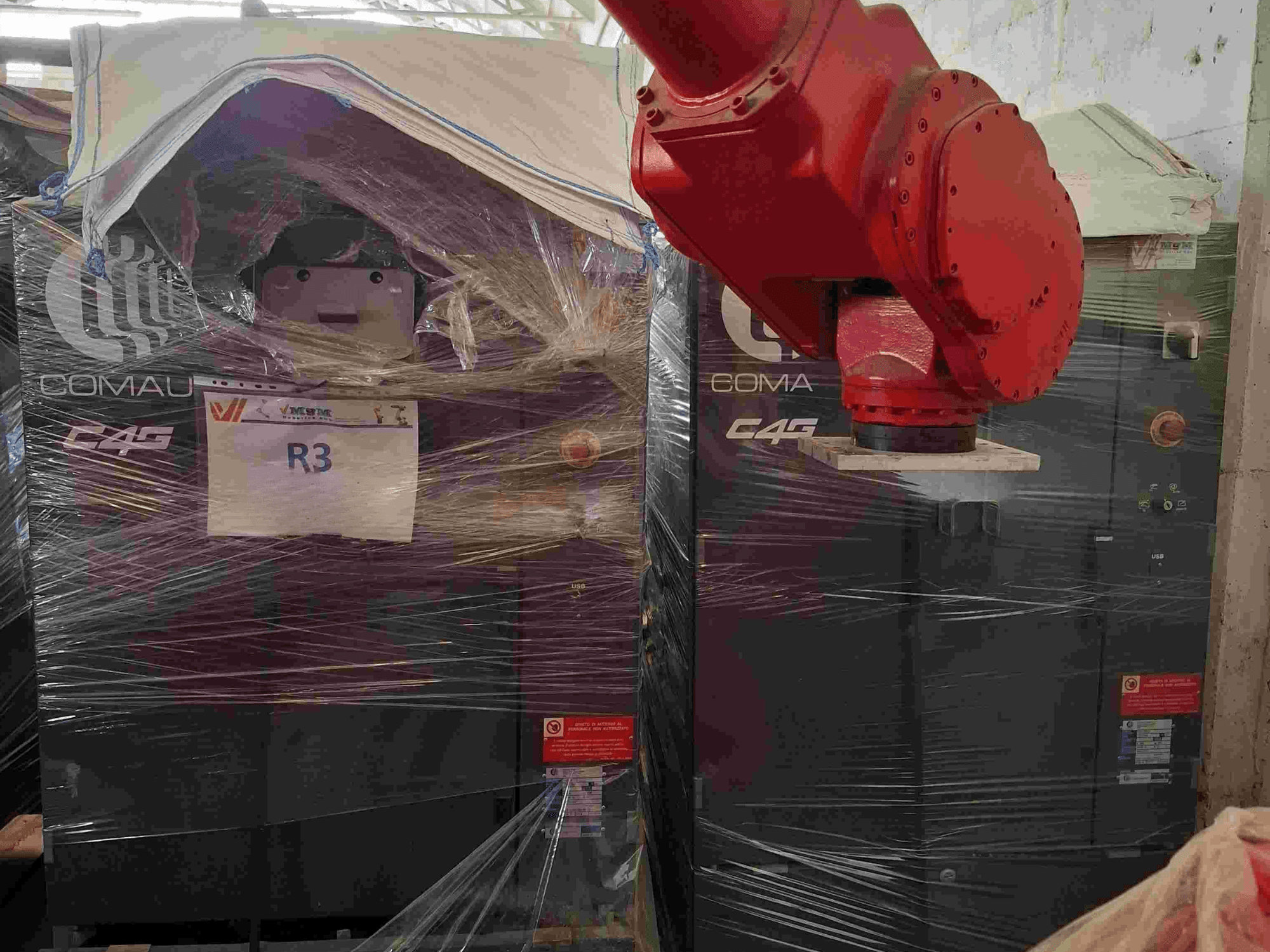 Robotické rameno Comau C4G zepředu, zabalené v plastu, označené "R3", s viditelnými varovnými nálepkami na stroji.