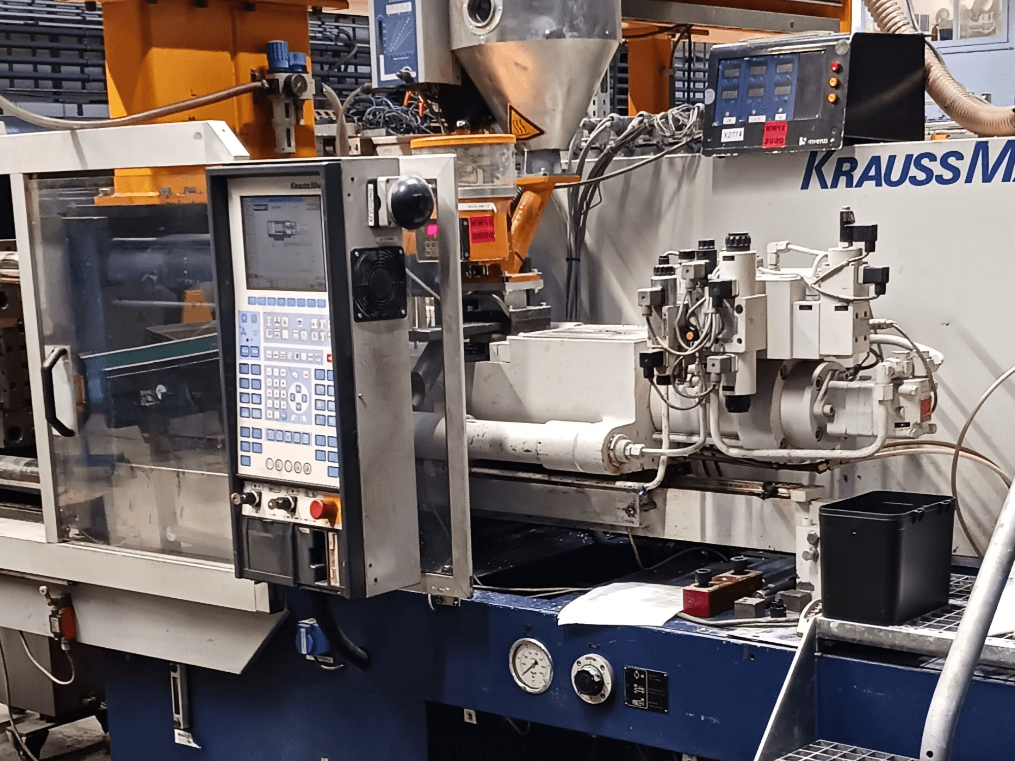 Vstřikovací stroj KraussMaffei s ovládacím panelem, hydraulickými jednotkami a nástroji v popředí, pohled zepředu.