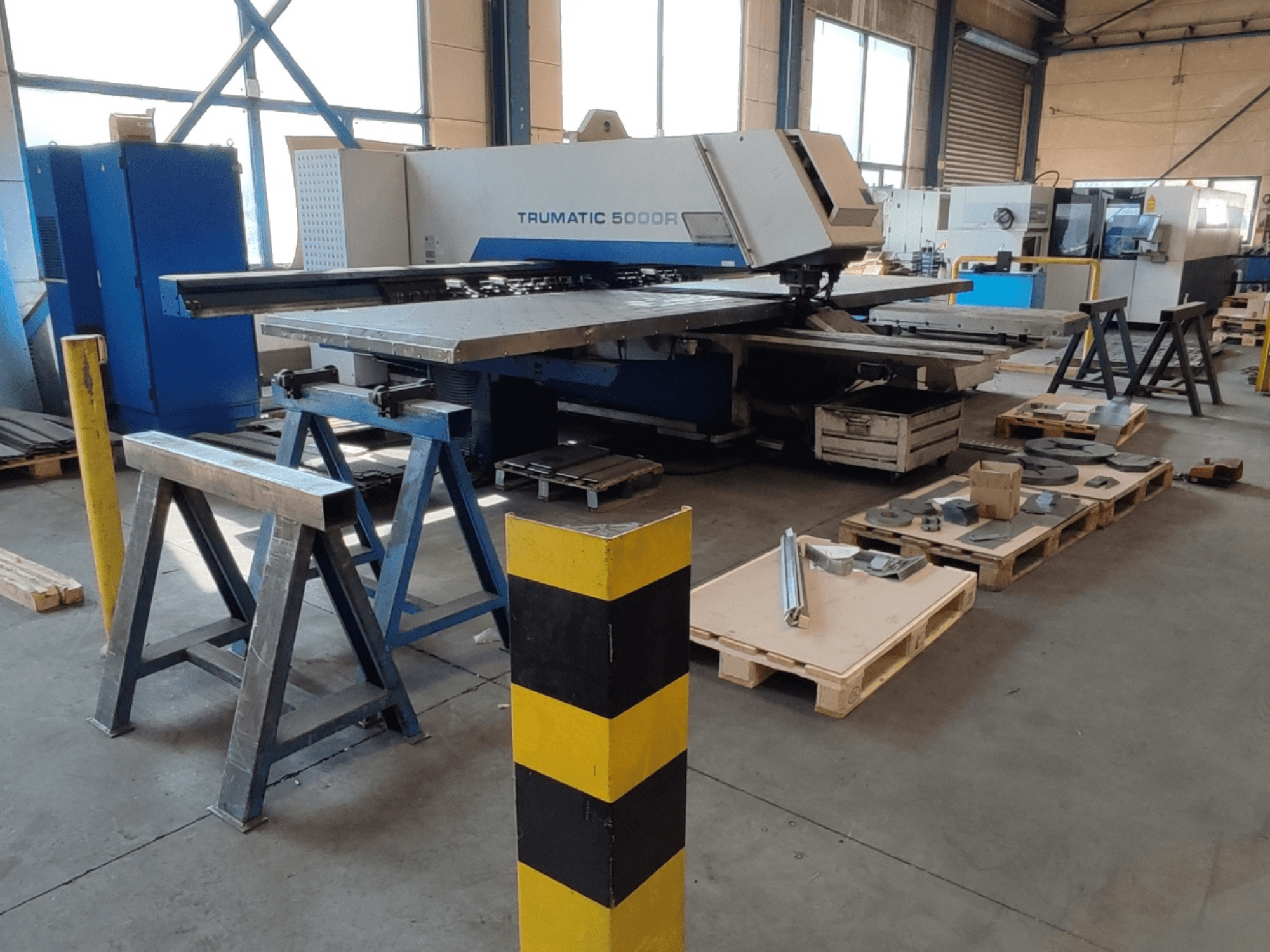 CNC děrovací stroj TRUMPF Trumatic 5000 R v dílně, pohled z boku s nástroji a materiály na paletách v blízkosti.