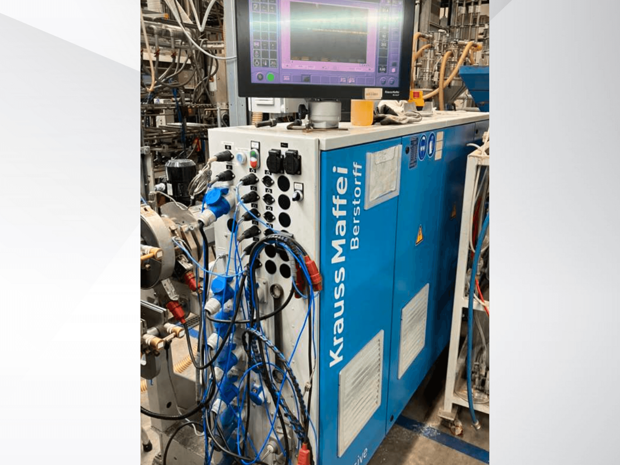 Průmyslový stroj Krauss Maffei Berstorff s ovládacím panelem a monitorem, s různými tlačítky a připojenými hadicemi, pohled z boku.