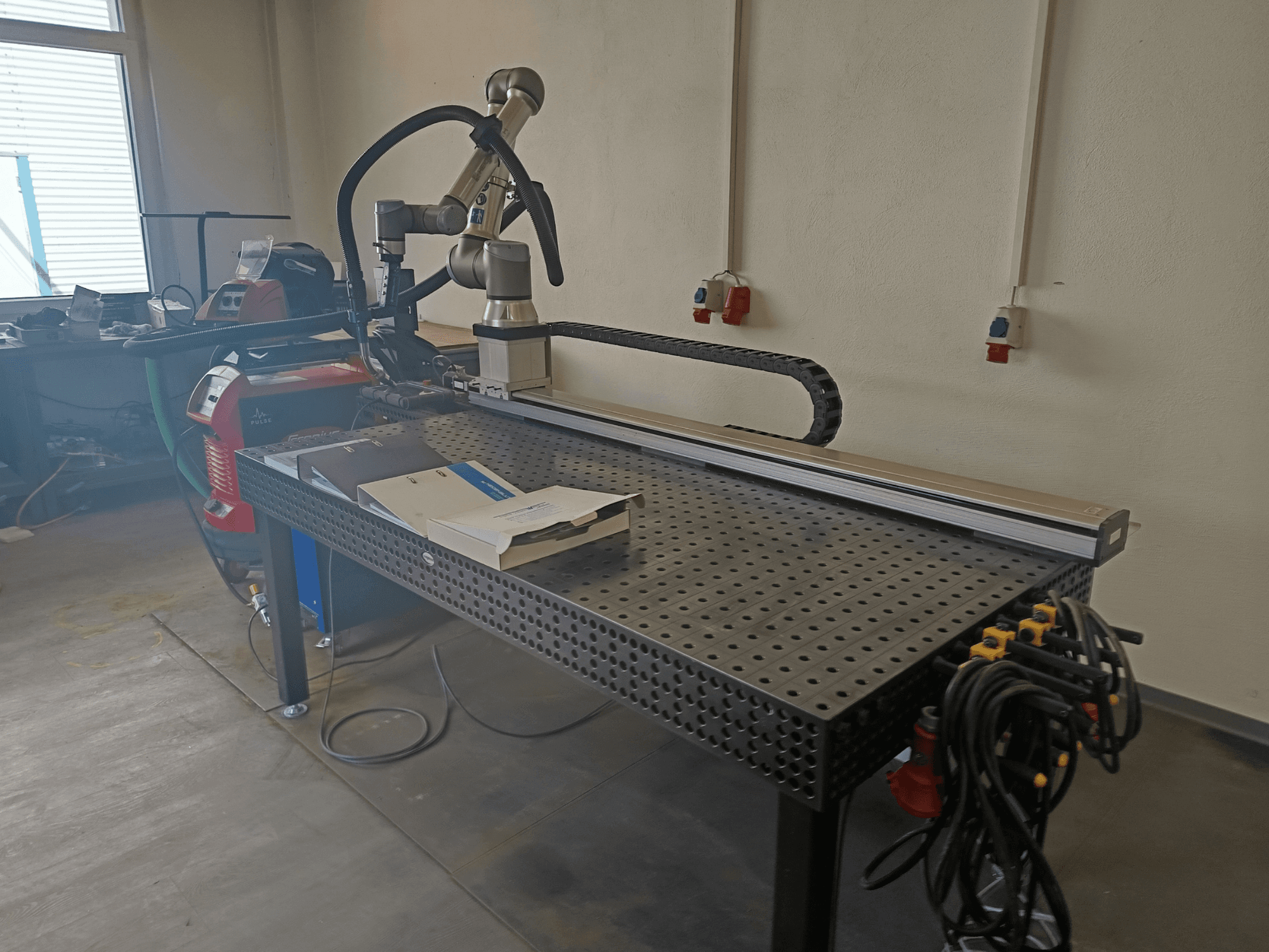 Svařovací buňka FRONIUS TransSteel 4000 Pulse při pohledu z boku, s robotickým ramenem, pracovním prostorem a napájecími přípojkami.