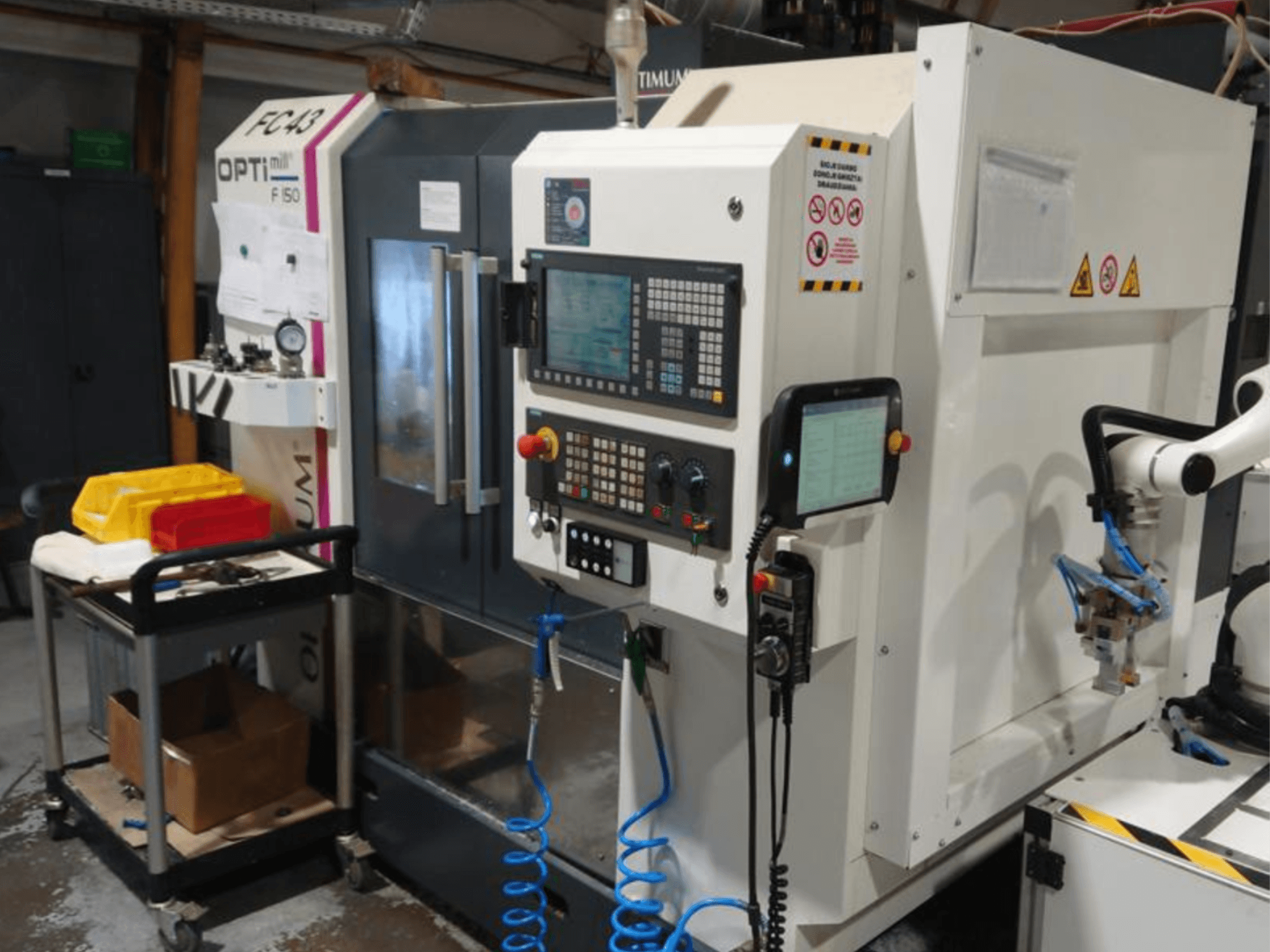 Ovládací panel frézky OPTIMUM F50 s hadicemi, nástroji a robotickým ramenem viditelný v dílně.