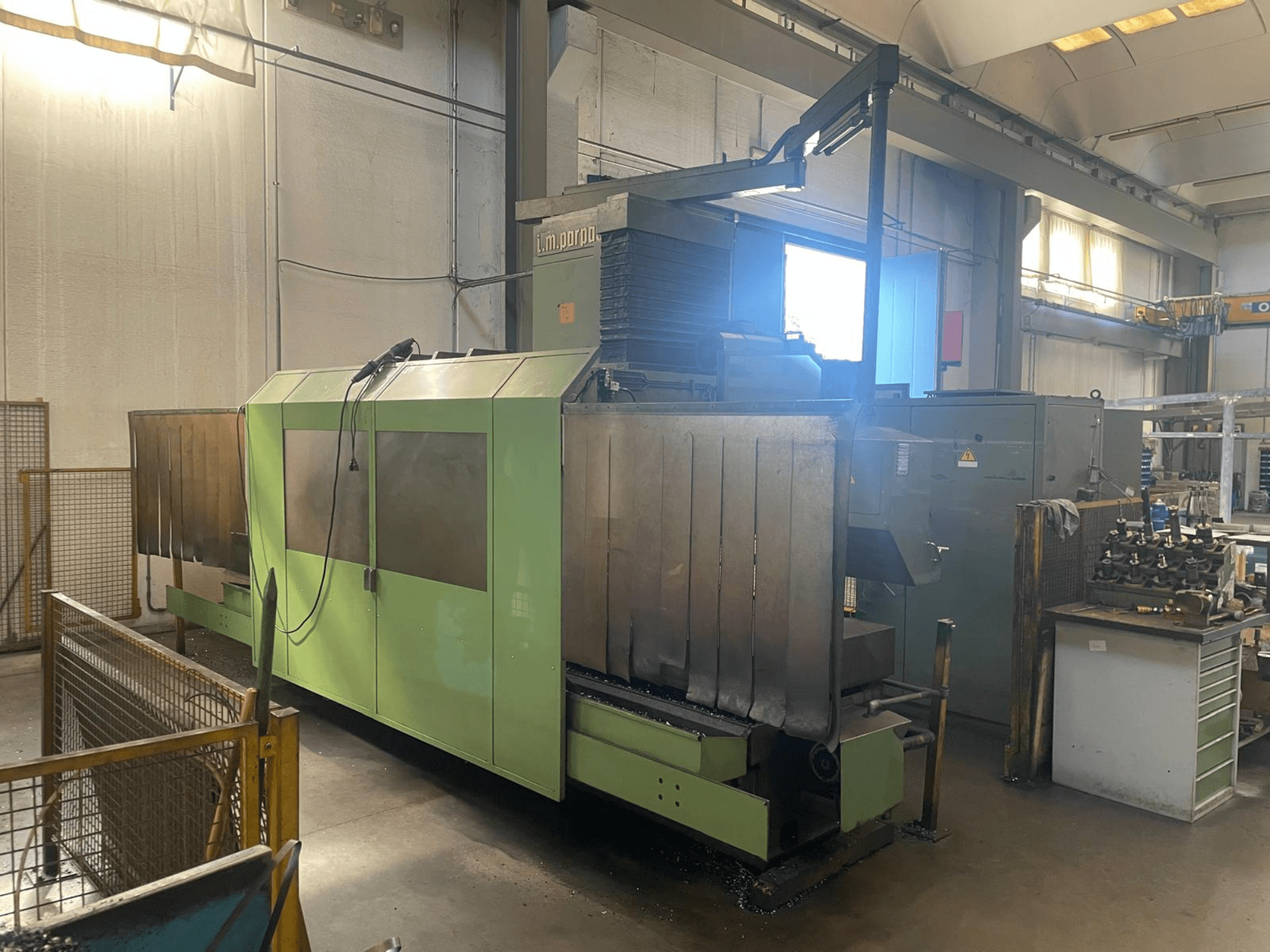 Zelený průmyslový stroj s viditelnými kovovými součástmi, pohled zepředu; s ovládacím panelem, bezpečnostními upozorněními a pracovním místem v blízkosti.