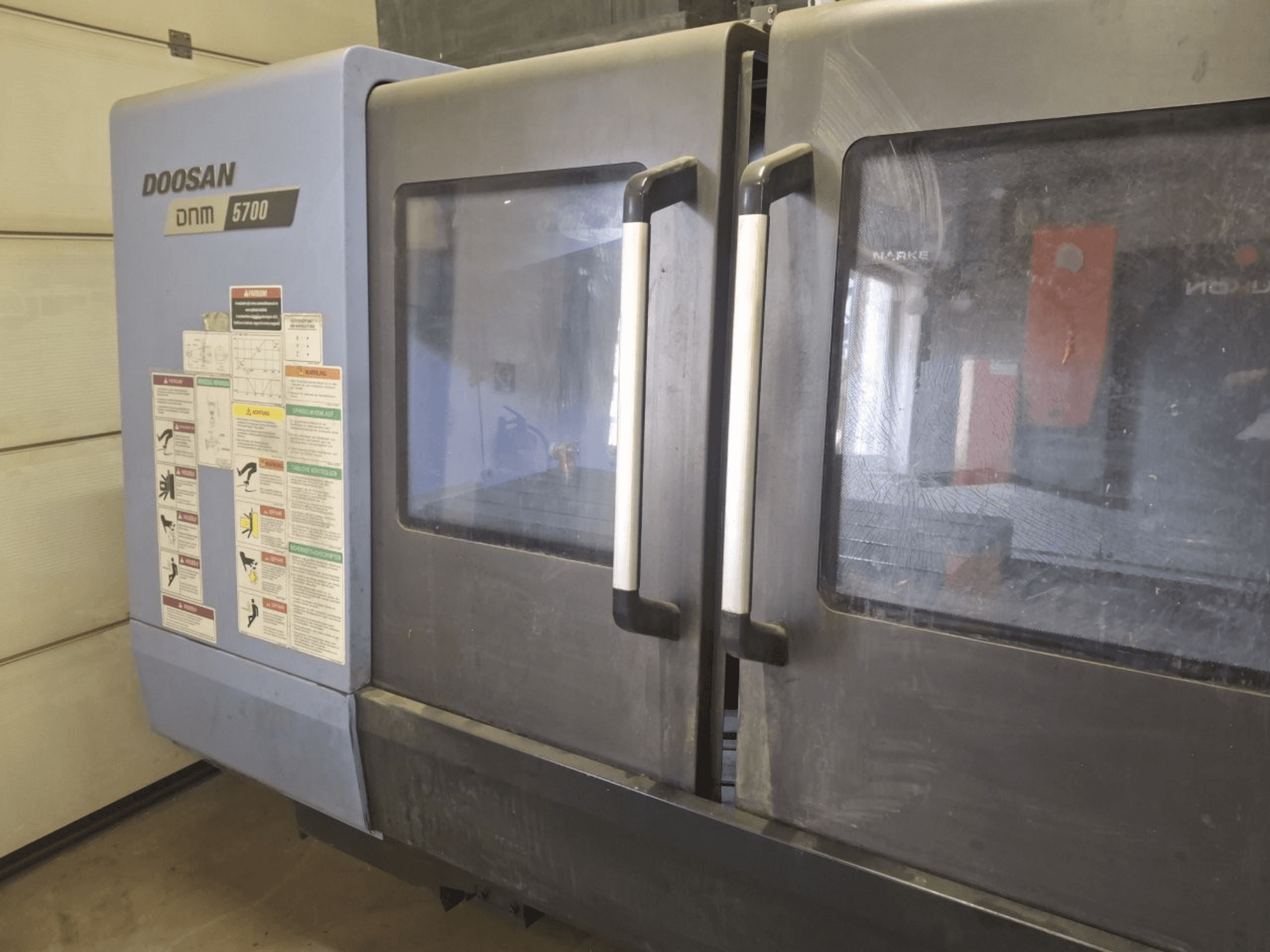Pohled zepředu na vertikální obráběcí centrum DOOSAN DNM 5700, na kterém jsou vidět dvoje dveře a ovládací panely s provozními pokyny.