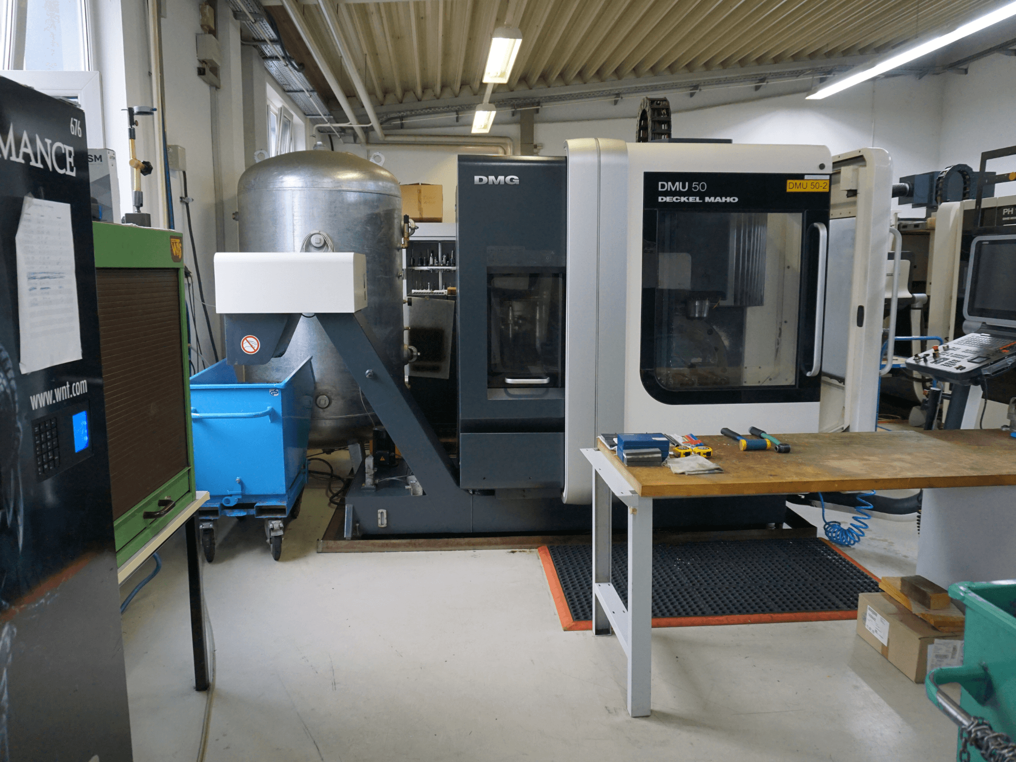 Frézka DMG DMU 50 v dílně, pohled zepředu, s nástroji na dřevěném stole a modrým košem na kolečkách poblíž.