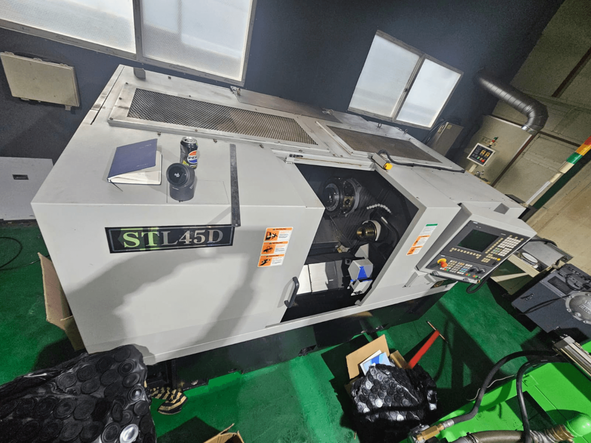 Stroj STL45D CNC při pohledu shora, zobrazující ovládací panel, válcový díl, notebook a plechovku nahoře.