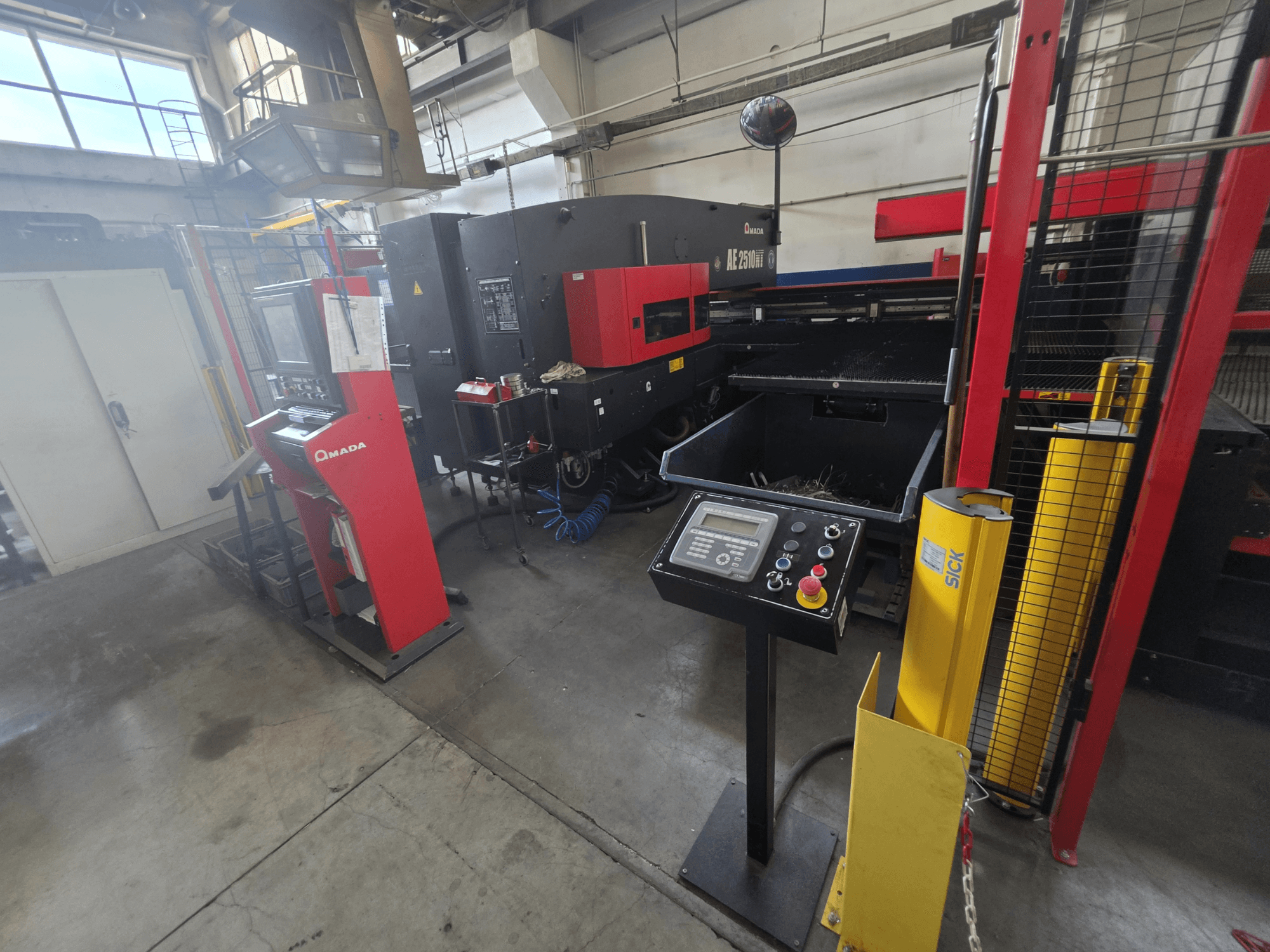 CNC děrovací stroj AMADA AE-2510NT, pohled zepředu s ovládacím panelem, bezpečnostními prvky a okolním průmyslovým prostředím.