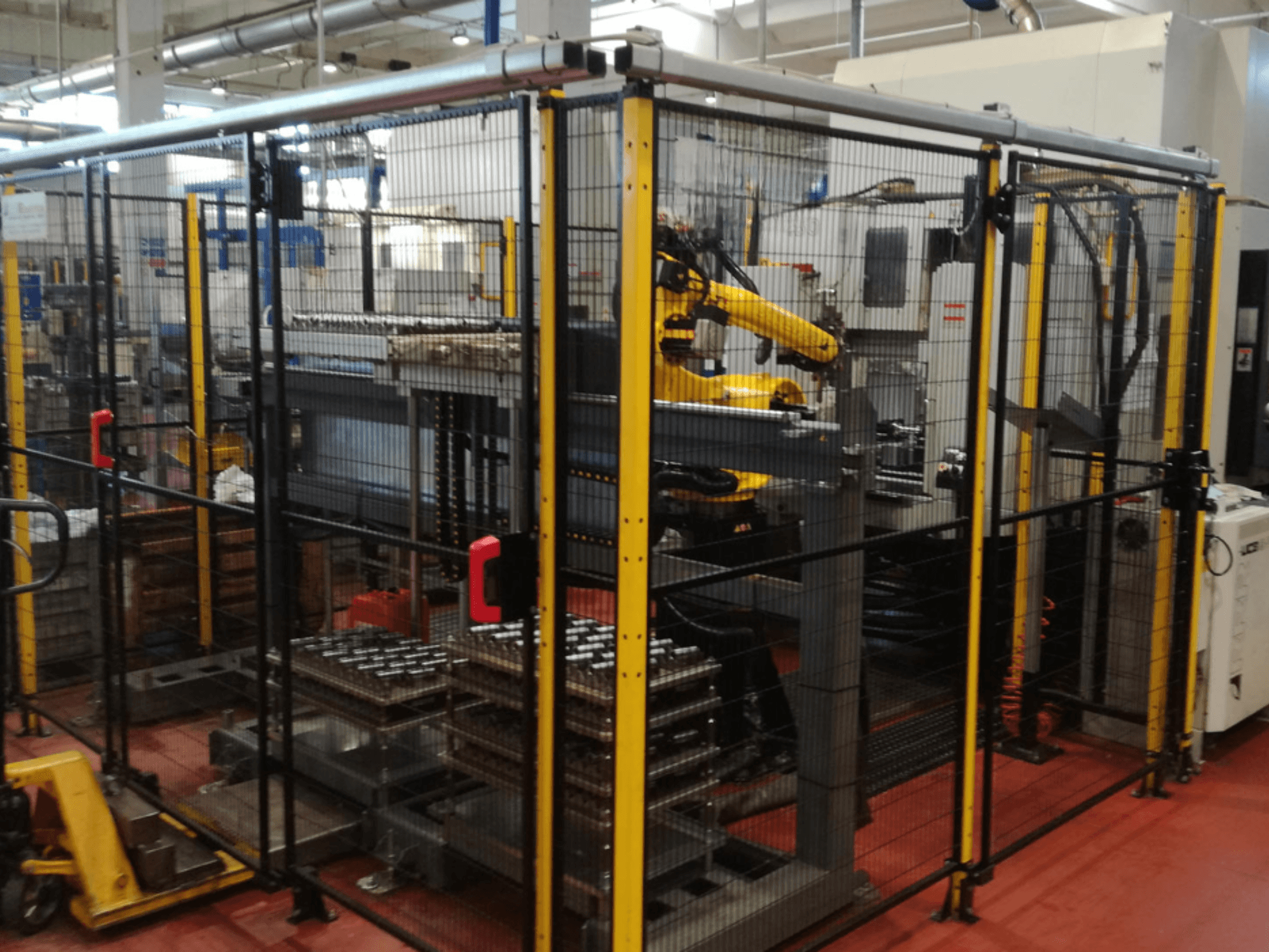 Vertikální obráběcí centrum Enshu EV450 v oploceném pracovním prostoru s robotickými rameny a několika na sobě uloženými obrobky. Pohled zepředu.