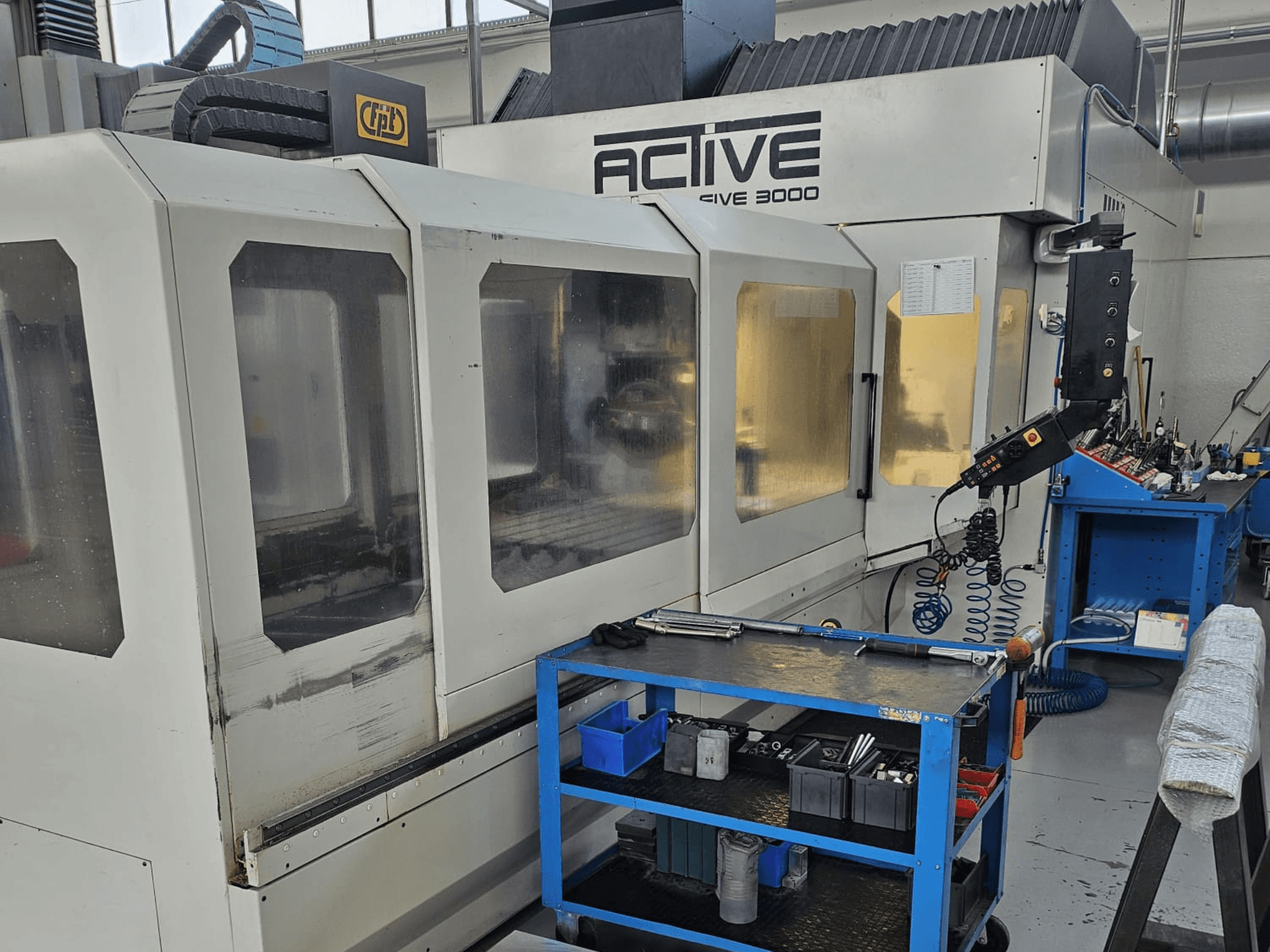 CNC stroj Active 3000 v dílně, pohled zepředu s nástroji na modrém vozíku, viditelné ovládací prvky a pracovní plocha uvnitř.