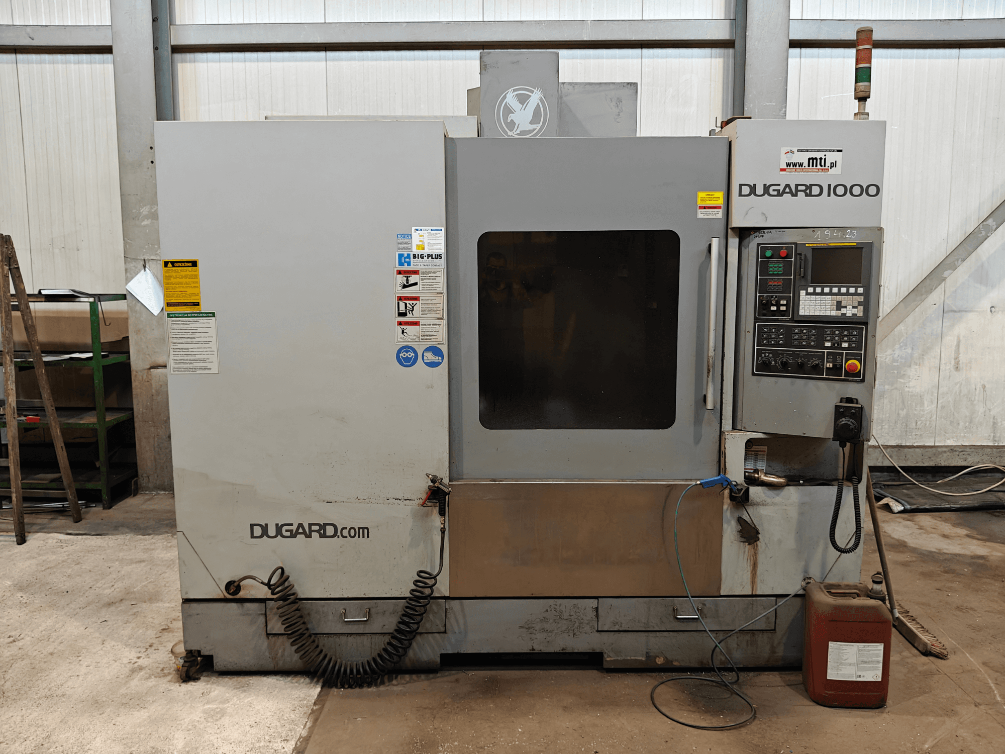 Stroj Dugard 1000 CNC, pohled zepředu, zobrazující ovládací panel, bezpečnostní štítky a nádobu na olej, v dílenském prostředí.