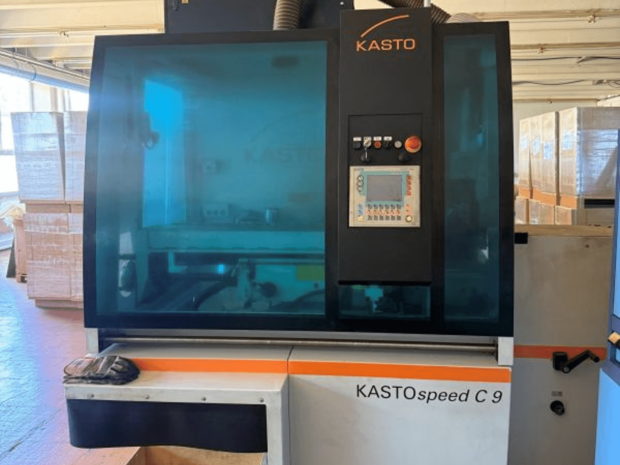 Pohled zepředu na kotoučovou pilu na kov KASTO KASTOspeed C 9 s ovládacím panelem a bezpečnostním sklem, v pozadí jsou dřevěné bedny.